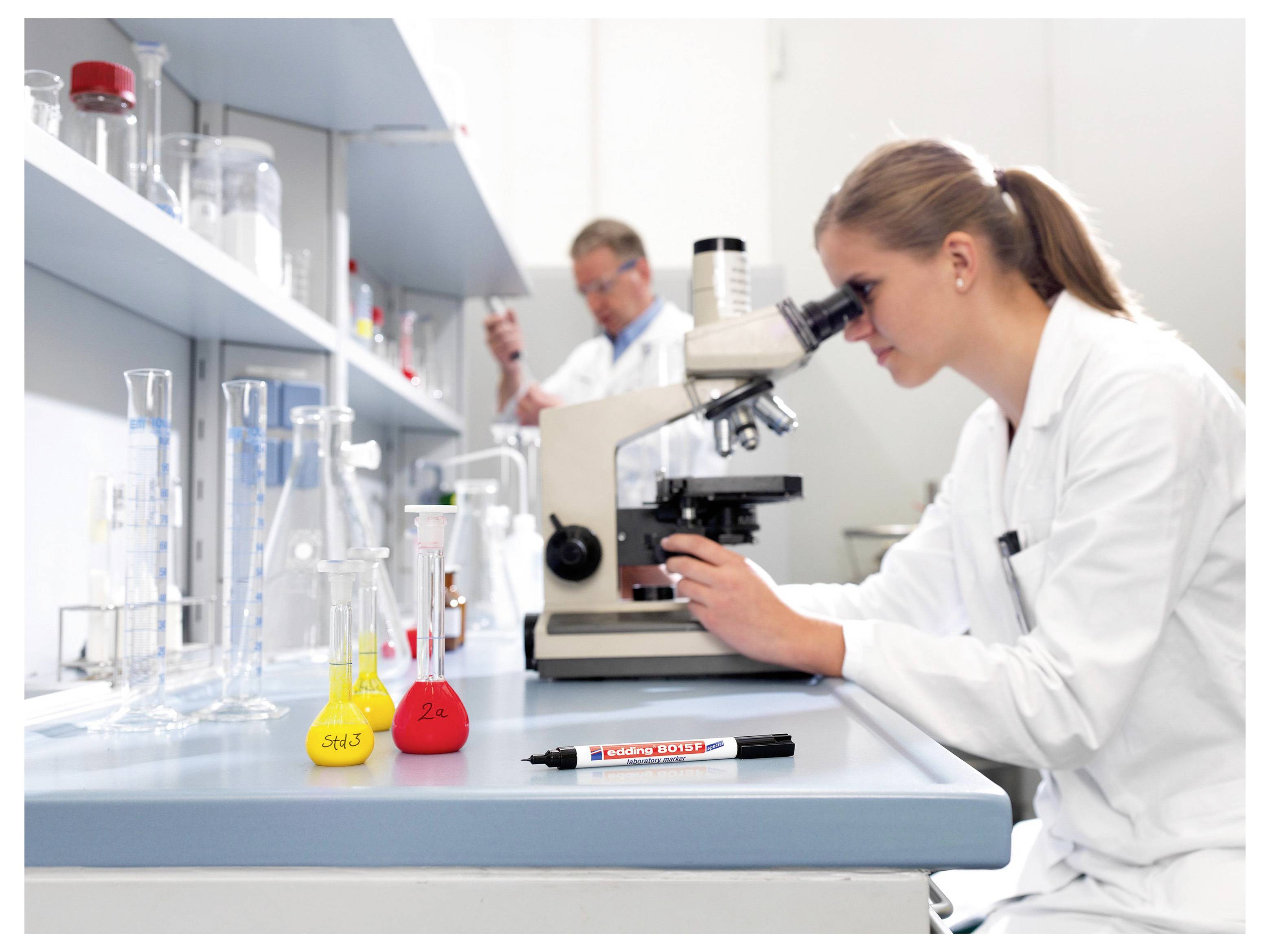Wissenschaftler in einem Labor, wobei eine Frau ein Mikroskop an einer Werkbank verwendet, die von Flaschen, Bechern und Chemikalien umgeben ist.