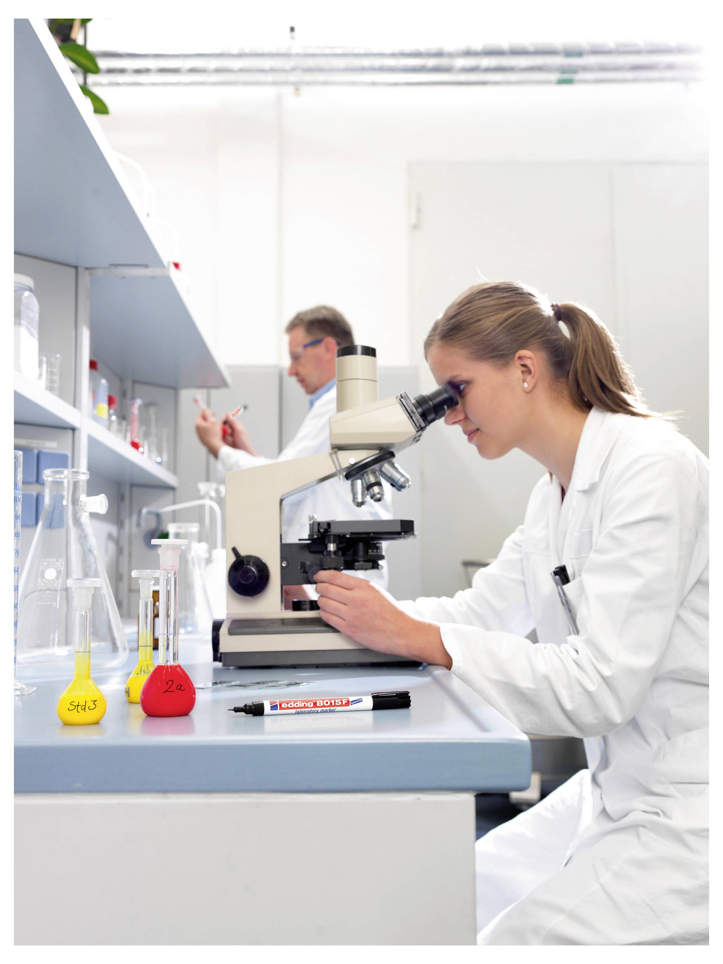 Wissenschaftler in einem Laborkittel schaut durch ein Mikroskop auf einer Laborbank mit Flaschen und farbiger Flüssigkeit, während ein anderer Forscher im Hintergrund arbeitet.