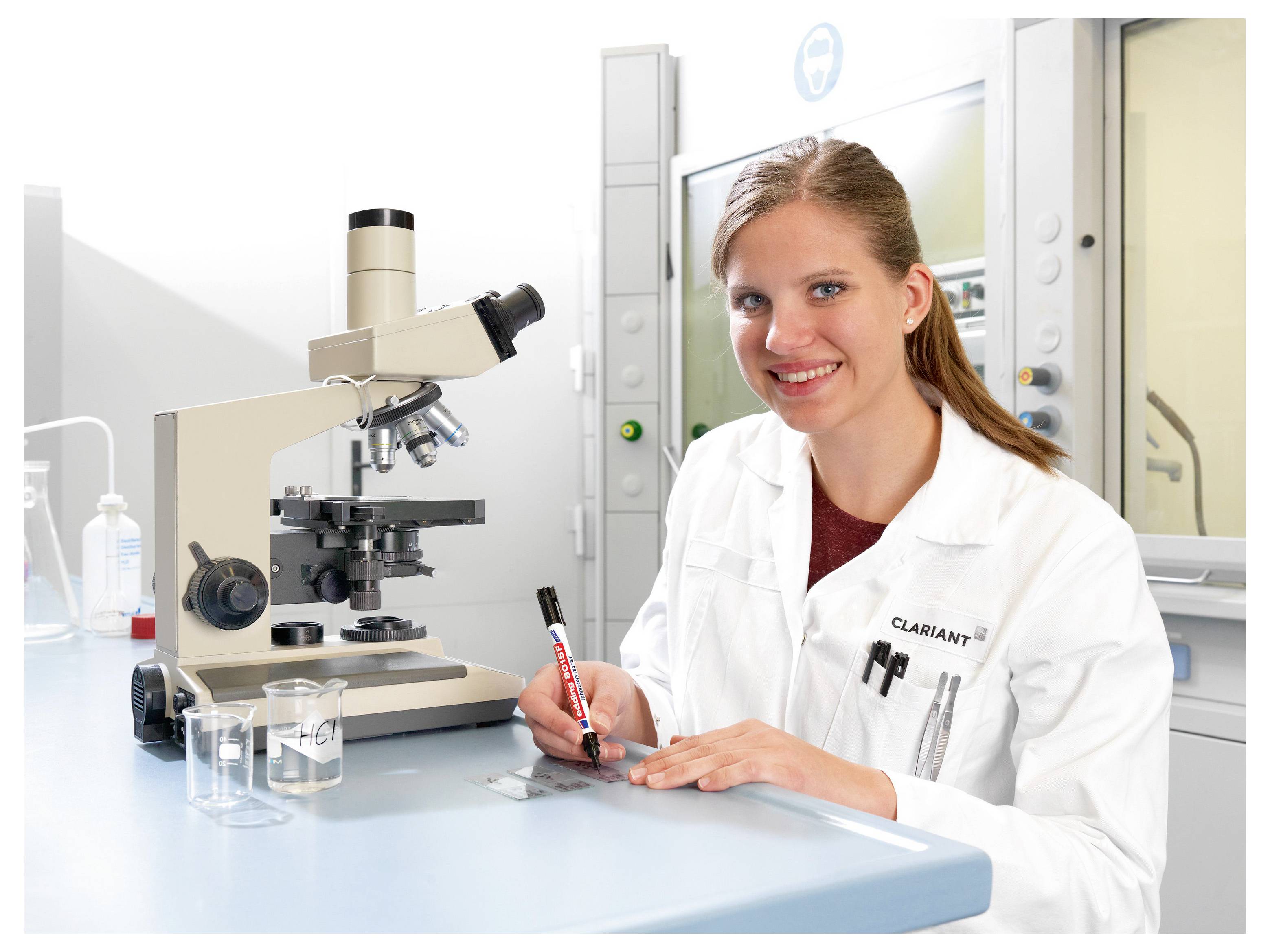 Lächelnde weibliche Wissenschaftlerin in einem Laborkittel schreibt an einer Laborbank neben einem Mikroskop und Glasbechern in einem hellen Labor.