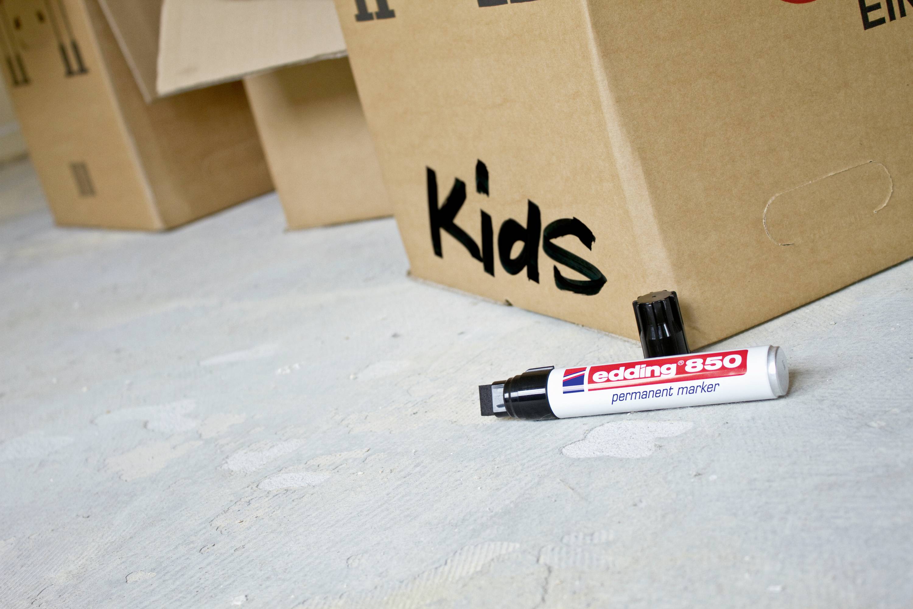 Kartons auf einem Boden, einer beschriftet mit 'Kids'. Ein Edding 850 Permanentmarker liegt davor.