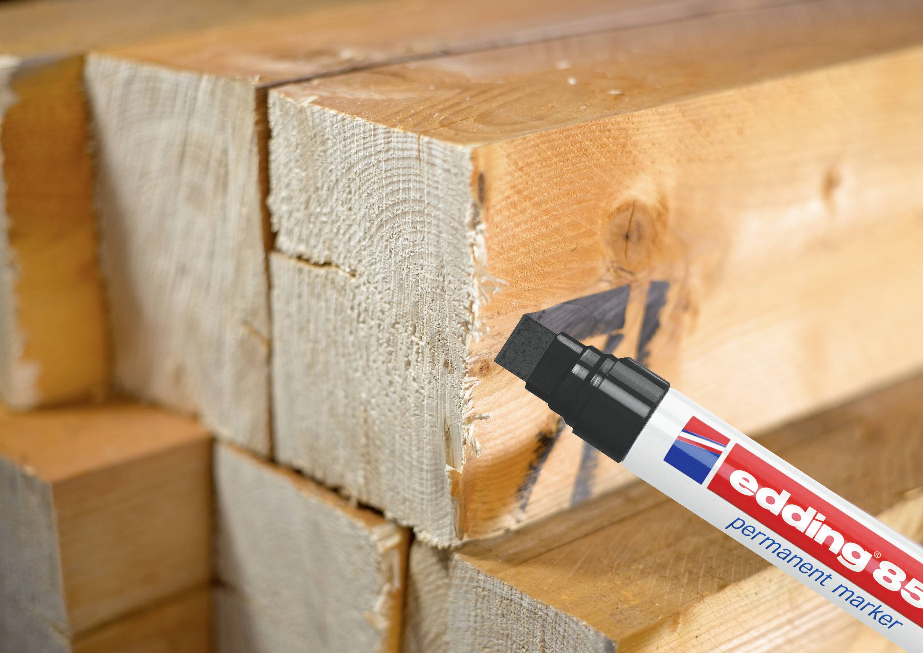 Ein schwarzer Permanentmarker beschriftet eine Holzbalkenoberfläche. Stapel von Holzlatten sind im Hintergrund sichtbar.