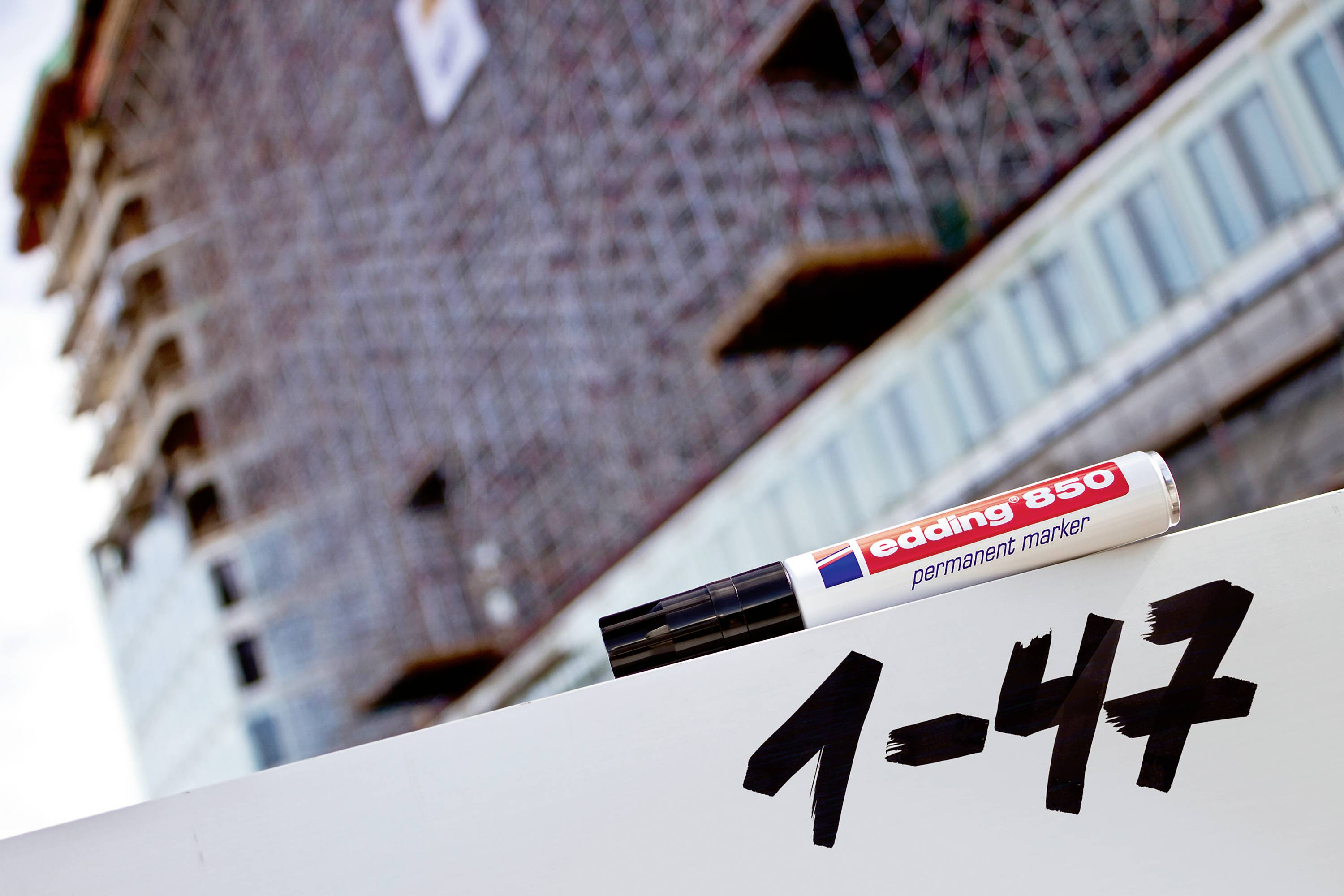 Ein 'edding 850' Permanentmarker liegt auf weißer Oberfläche, mit schwarzer Schrift '1-47'. Im Hintergrund ist ein Gebäude im Bau.