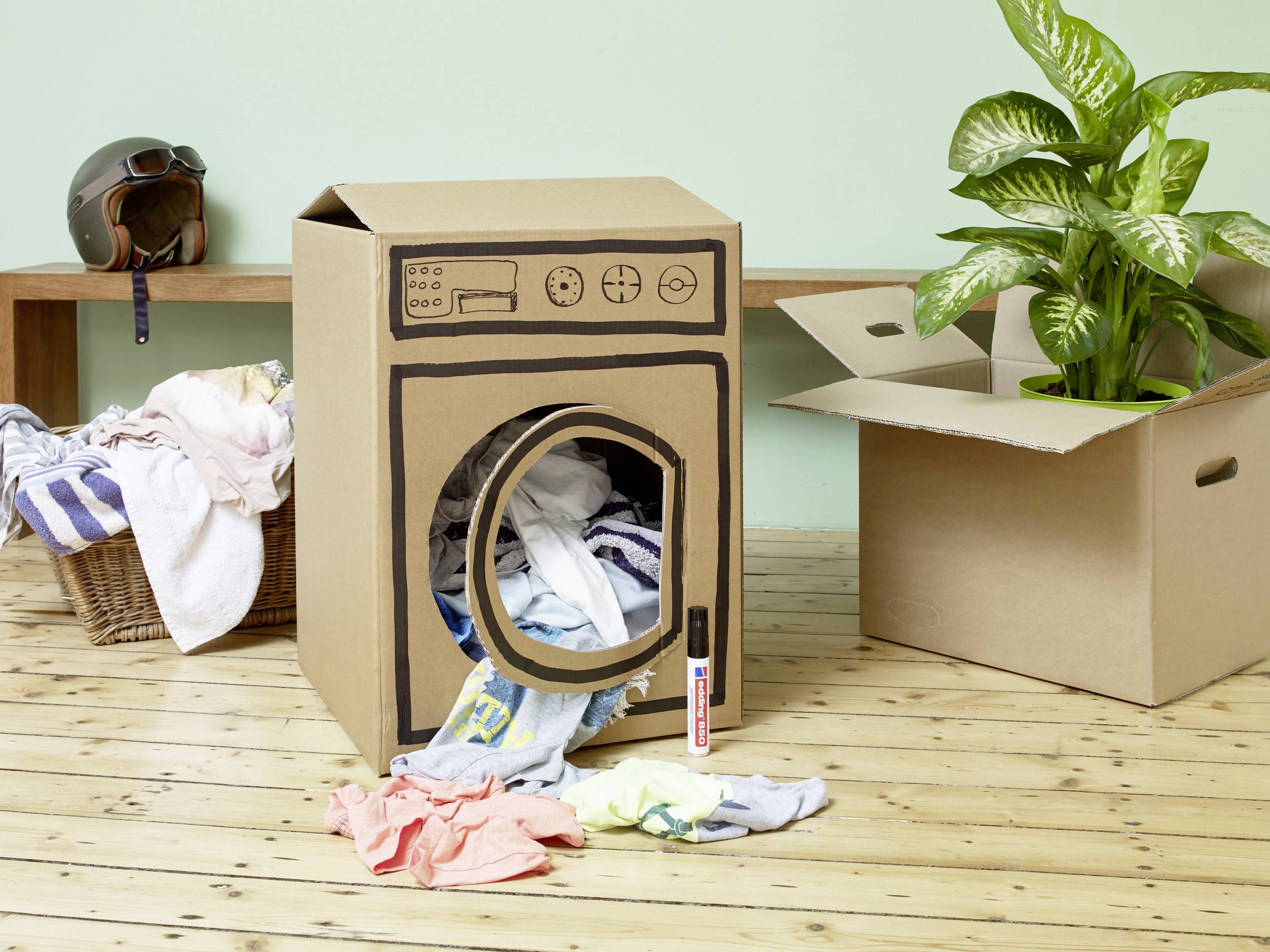 Ein aus Karton gebastelter Spiel-Wäschetrockner steht auf einem Holzboden. Daneben liegen und hängen bunte Kleidungsstücke.