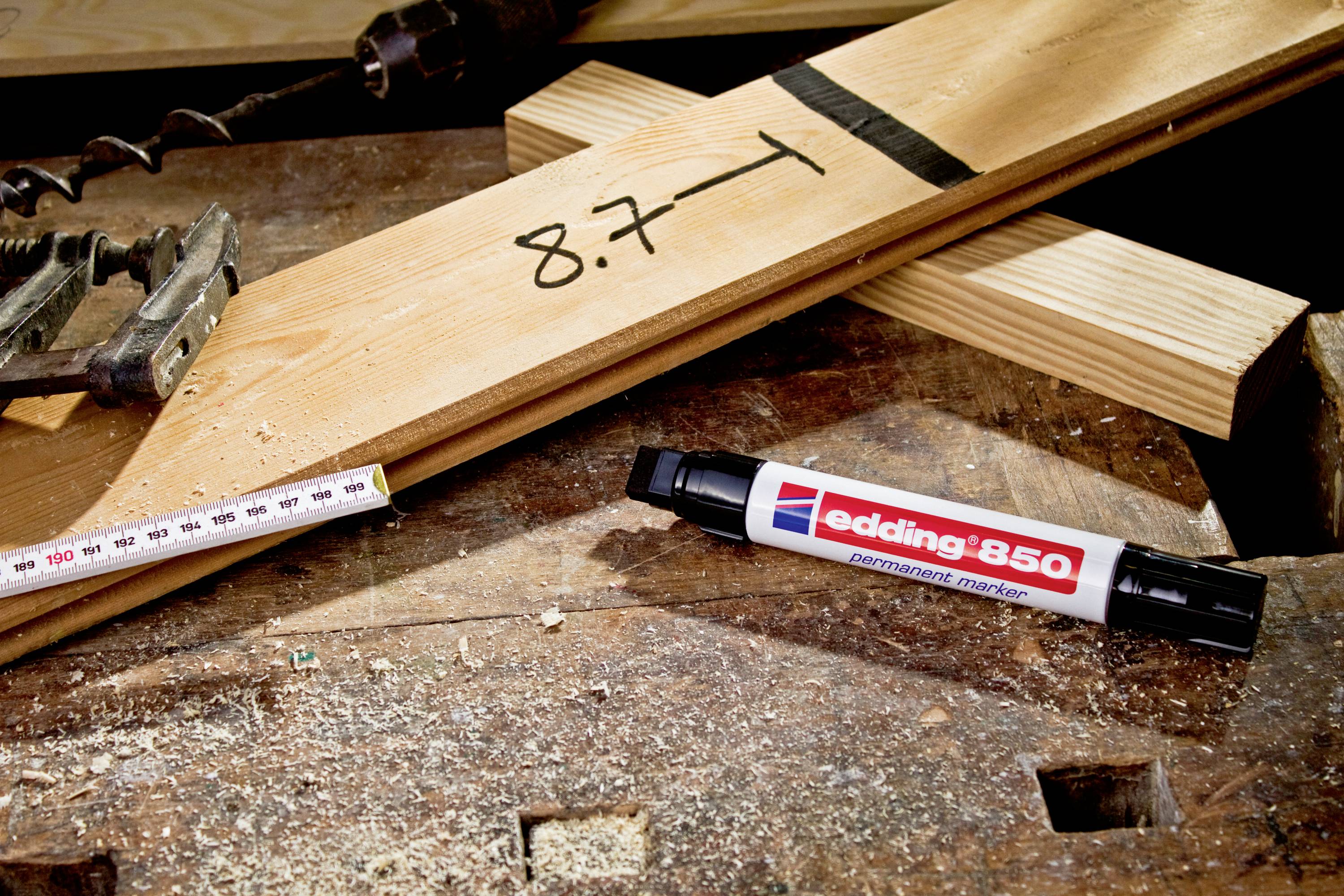 Ein Holzbrett mit der handschriftlichen Notiz '8.7', daneben ein Maßband und ein schwarzer Permanentmarker auf einer Werkbank.