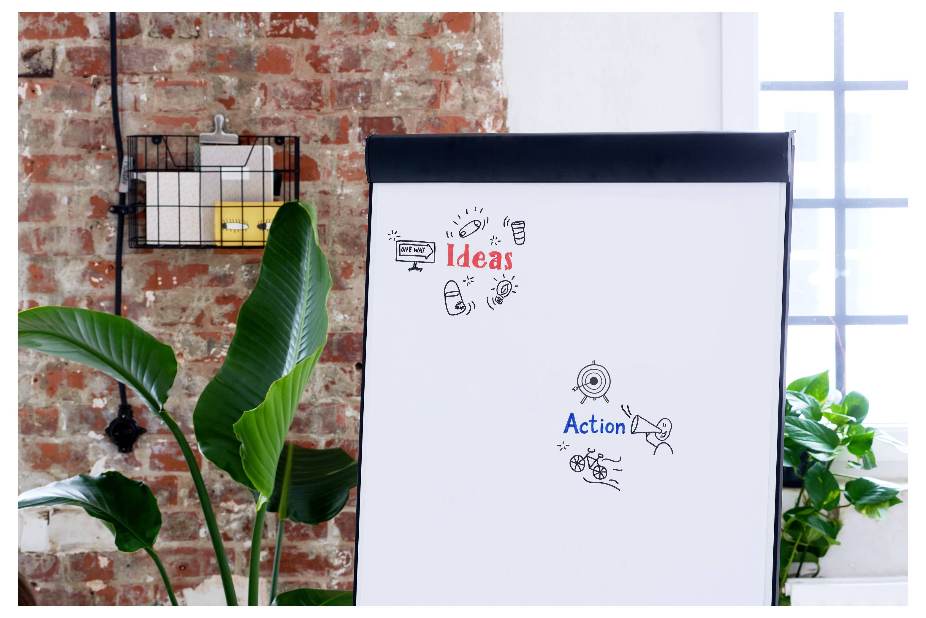Flipchart mit den Beschriftungen „Ideen" und „Maßnahmen" mit Doodle-Symbolen, in einem hellen Bürobesprechungsraum mit Pflanzen und sichtbarer Backsteinwand.