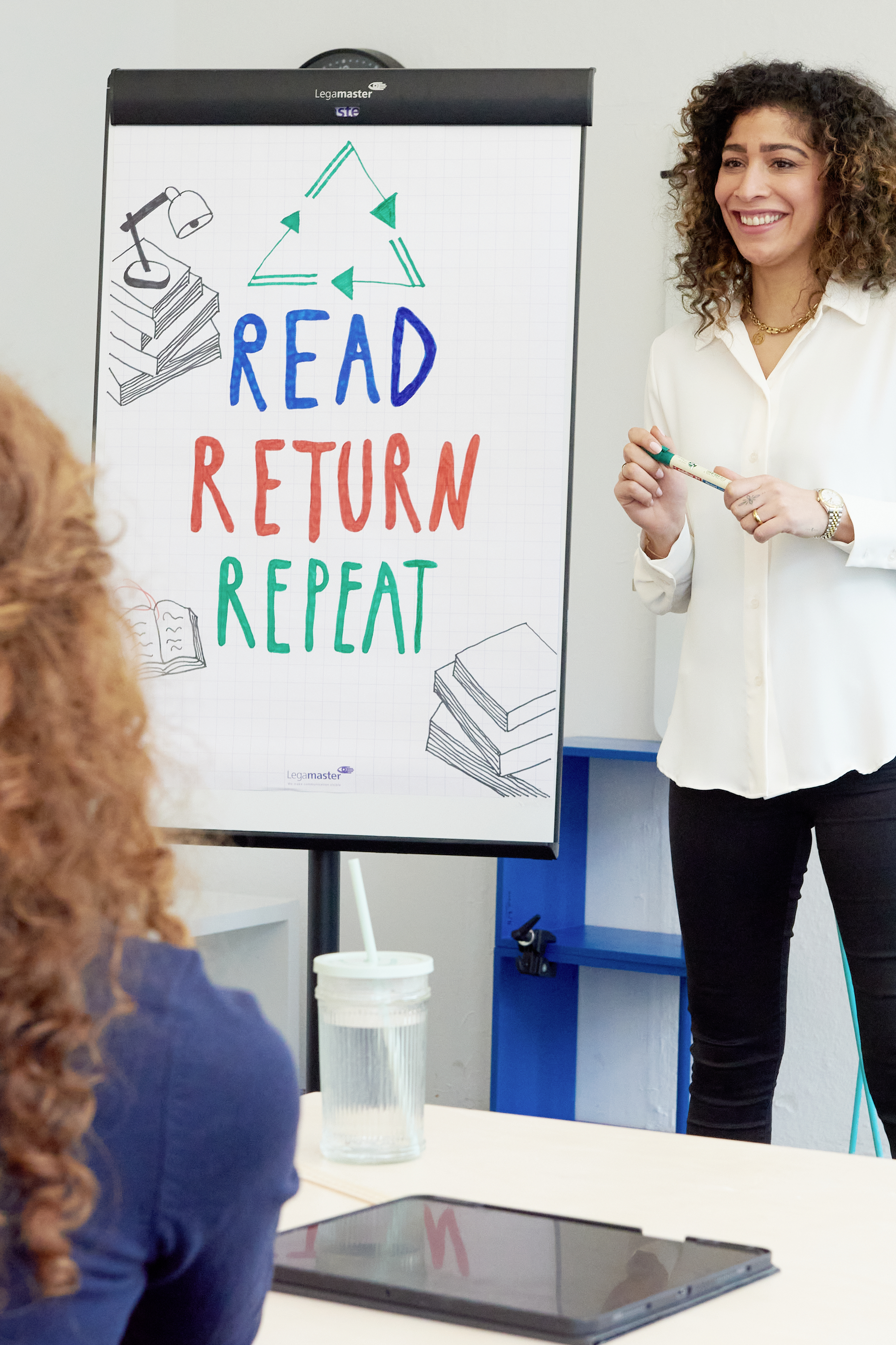 Eine Frau steht neben einem Flipchart mit der Aufschrift 'Read Return Repeat' inmitten einer Besprechung mit einer sitzenden Person.