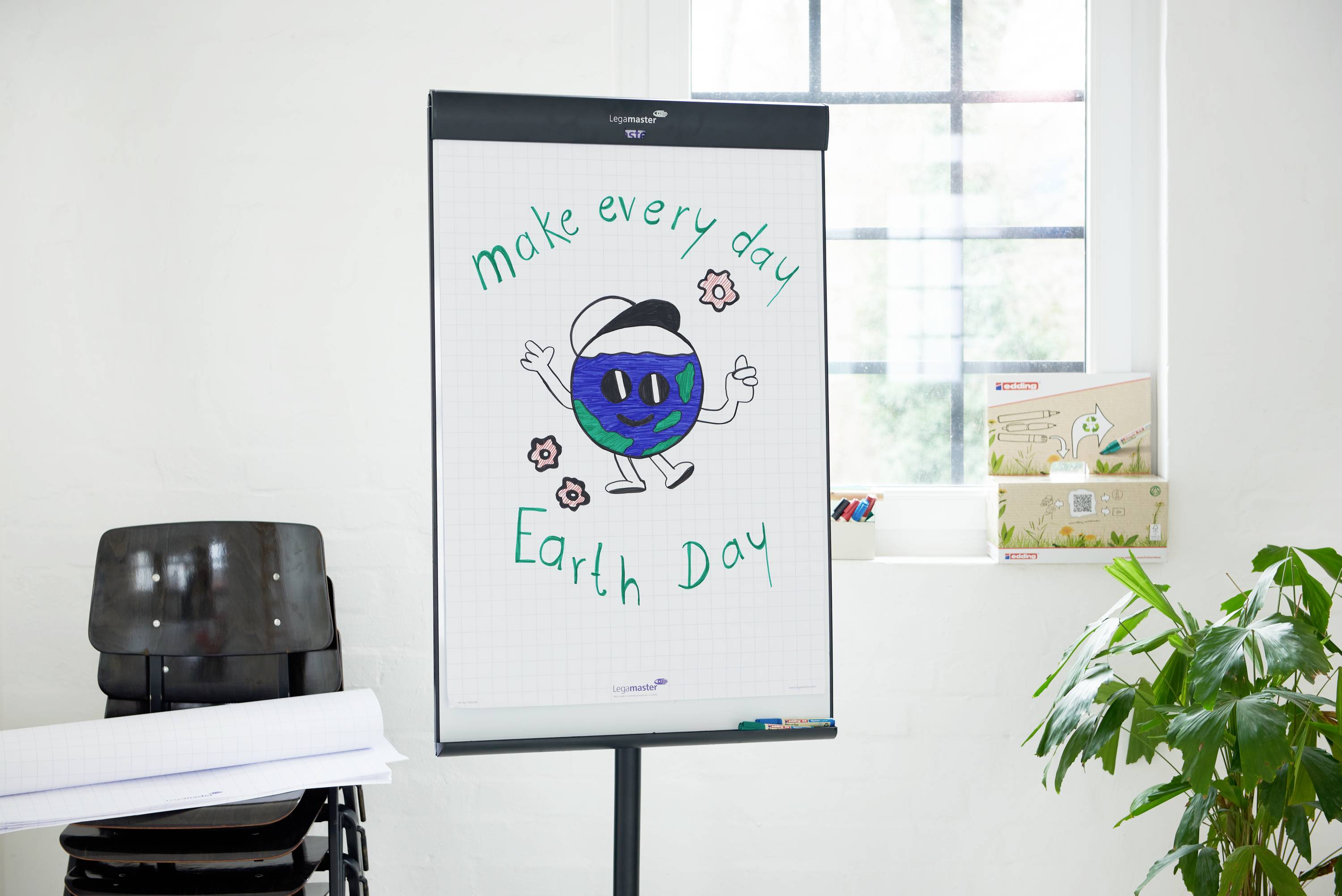 Flipchart mit der Zeichnung eines lachenden Planeten mit Armen und Beinen, umgeben von Blumen und dem Text 'make every day Earth Day'. Raum mit Fenstern und Pflanzen im Hintergrund.