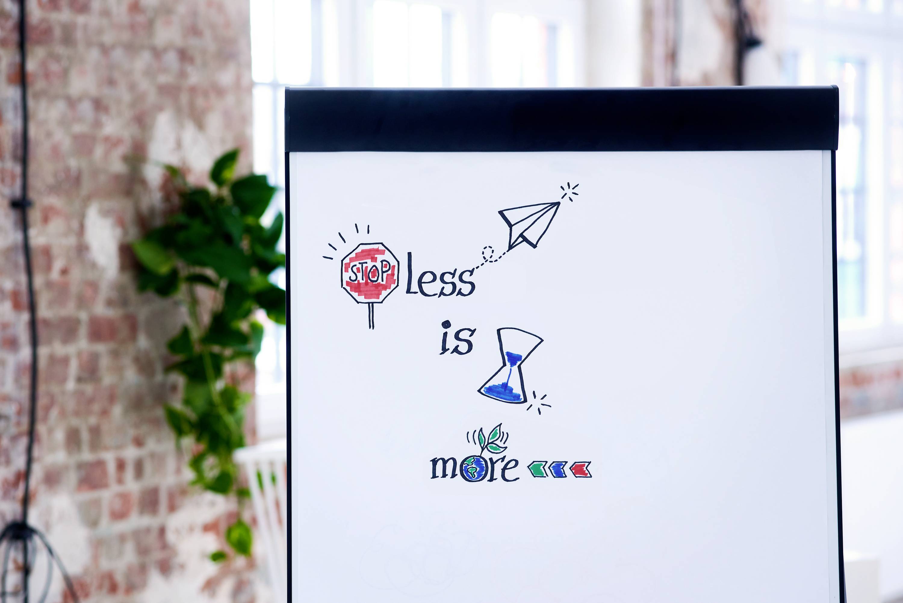 Ein Whiteboard zeigt die Inschrift 'Stop Less is More' mit Symbolen: ein Stoppschild, Papierflieger, Sanduhr und Pflanzen. Hintergrund ist ein Backsteinzimmer.