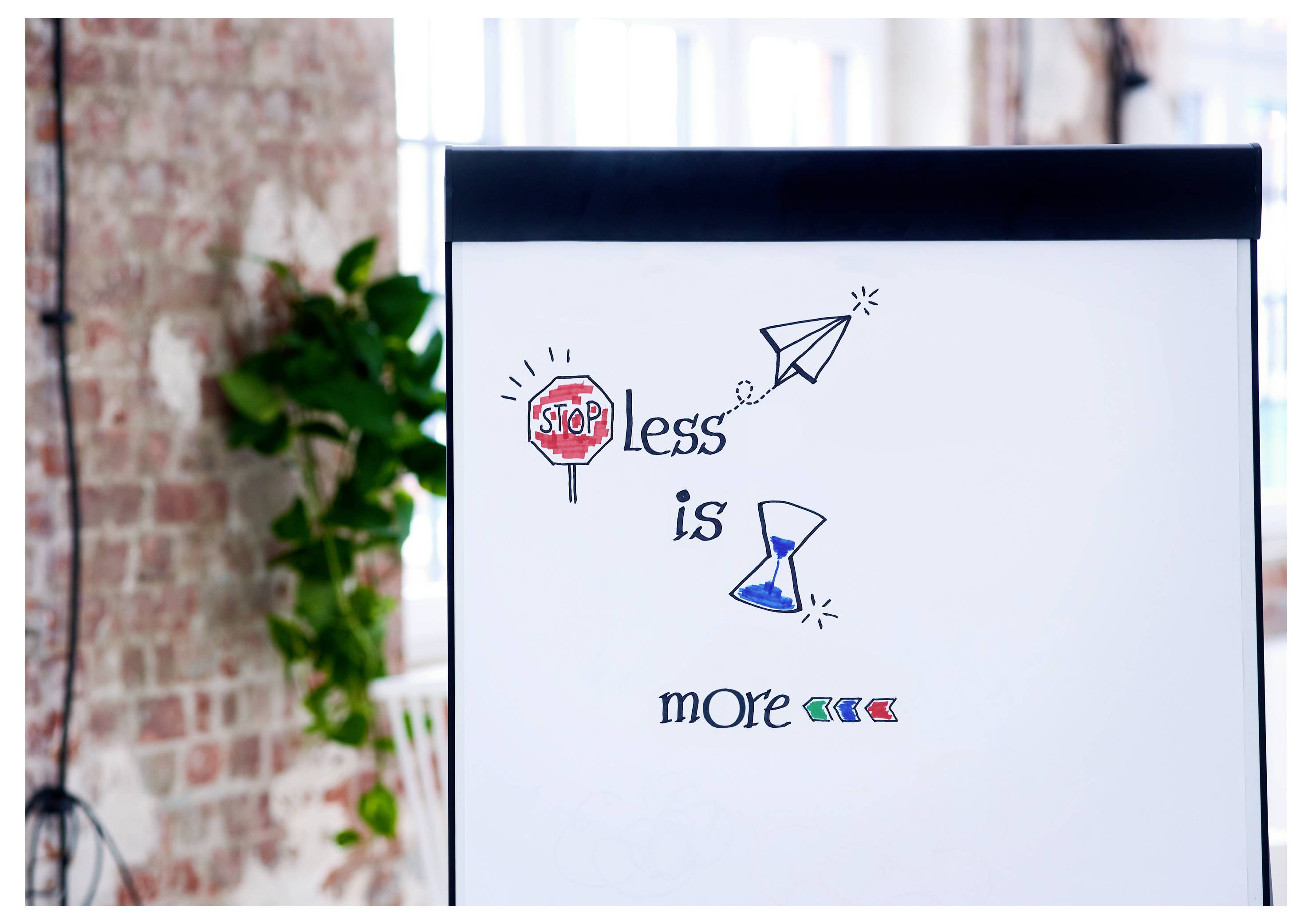 Flipchart mit dem Satz „Weniger ist mehr" illustriert durch ein Stoppschild, ein Papierflugzeug und eine Sanduhr in einer Büroumgebung.