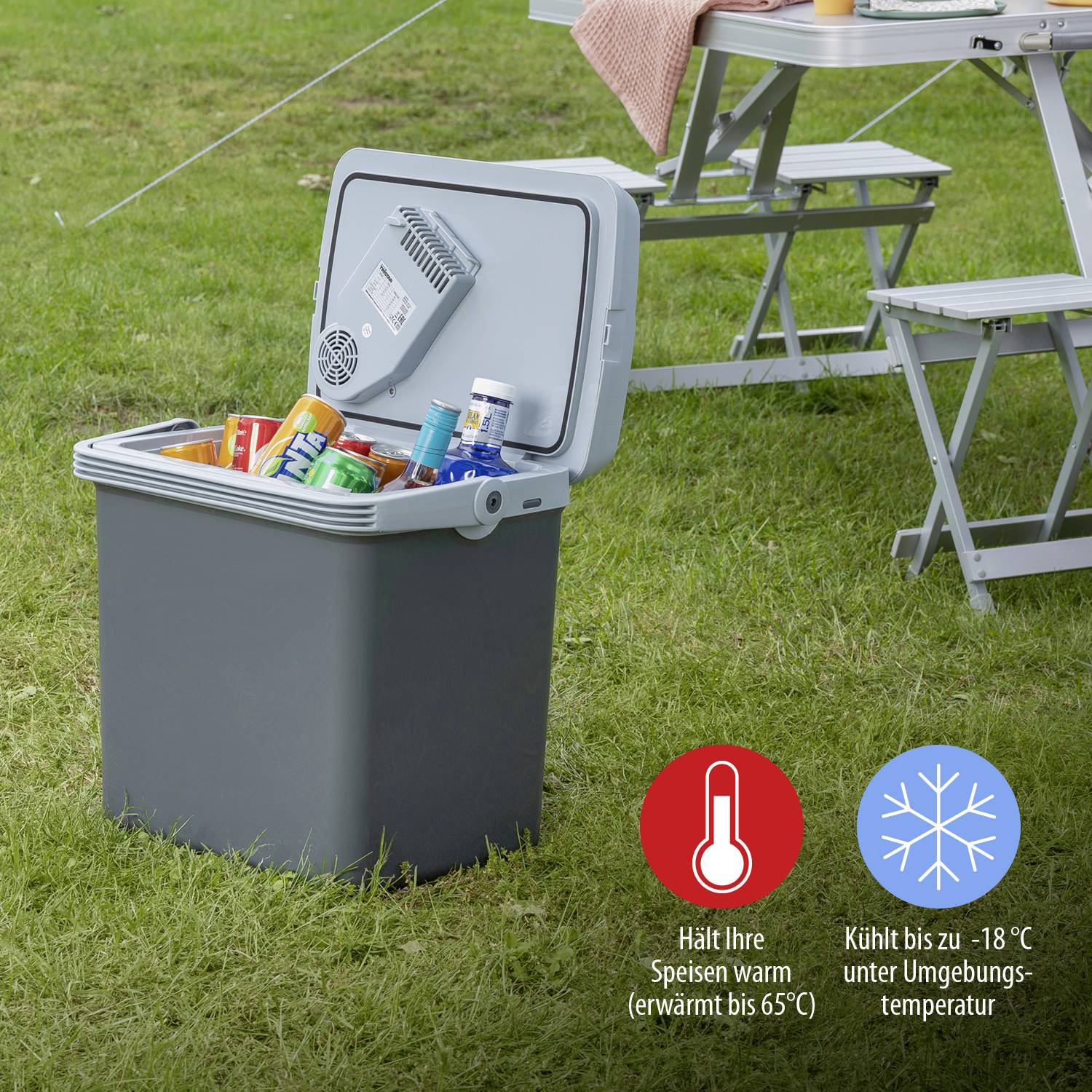 Kühlbox im Freien, geöffnet, gefüllt mit Getränken und Lebensmitteln, platziert neben einem Campingtisch und Stuhlset auf dem Rasen.