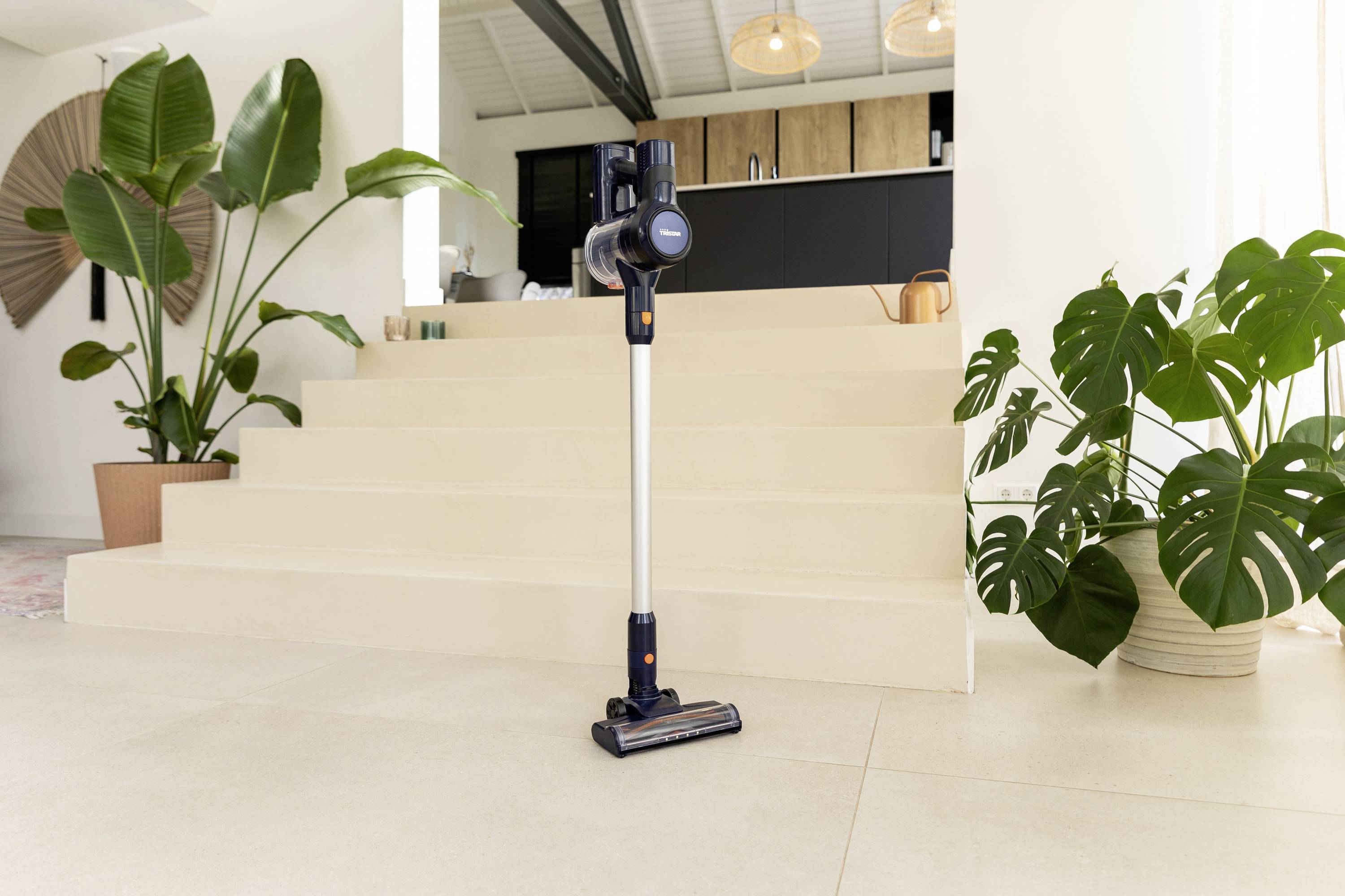 Kabelloser Staubsauger steht auf hellem Boden vor einer Treppe in einem modern eingerichteten Raum mit großen Zimmerpflanzen.