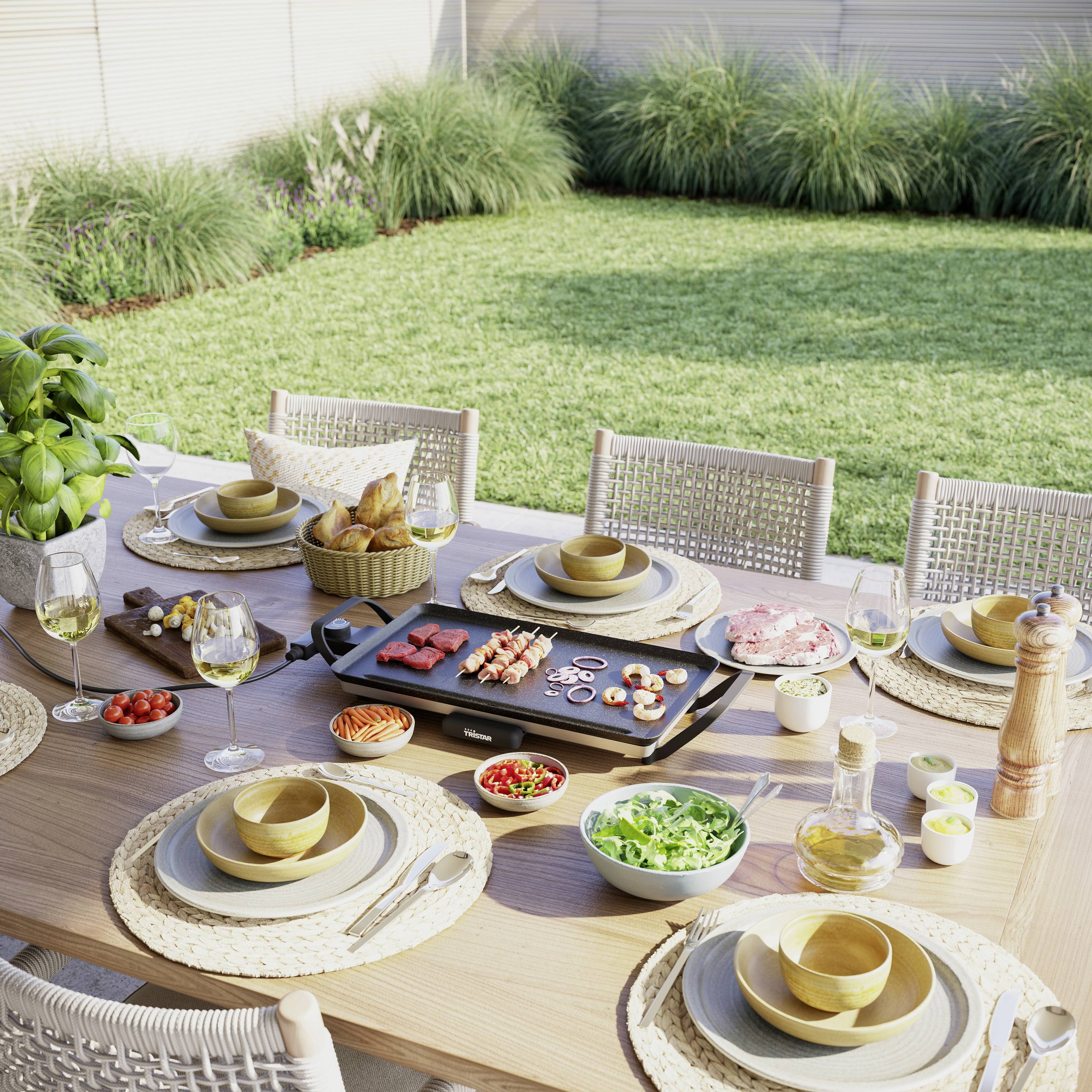 Ein gedeckter Gartentisch mit Grillplatte, auf der Fleisch und Gemüse zubereitet werden, umgeben von Geschirr und Gläsern. Im Hintergrund ein Rasen.