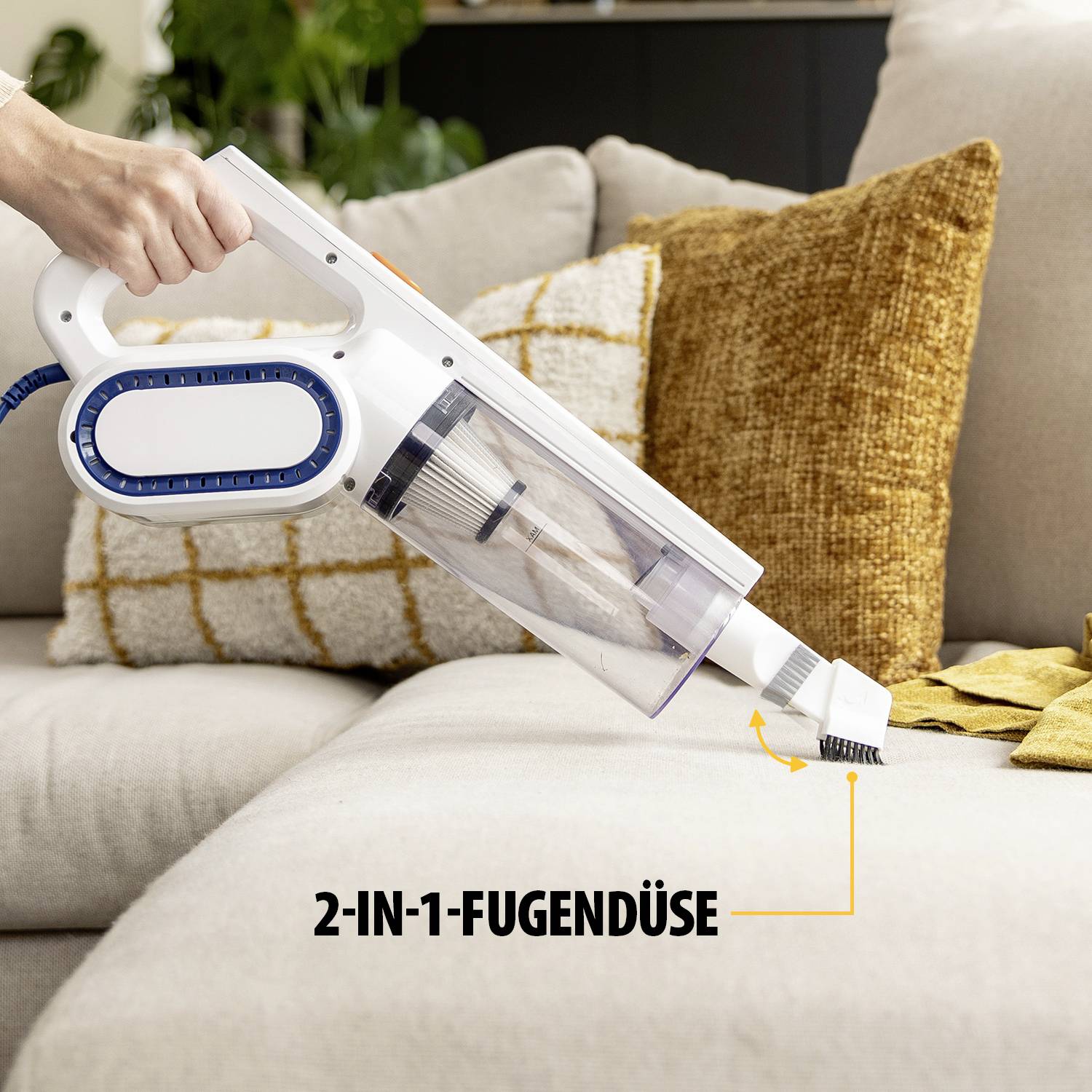 Eine Person benutzt einen tragbaren Staubsauger mit spezieller 2-in-1-Fugendüse, um eine Couch abzustauben. Im Hintergrund sind Kissen.