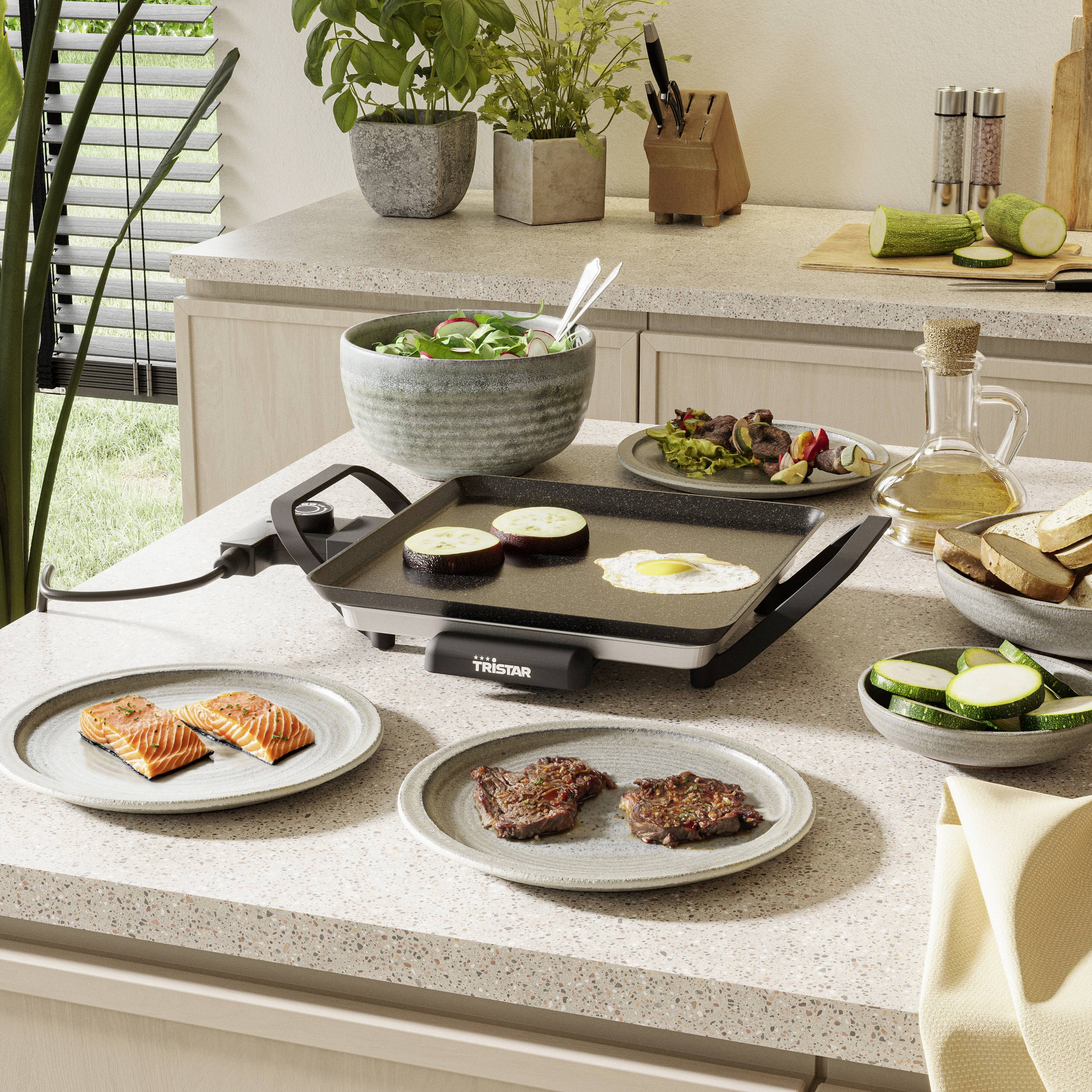 Eine Küche mit einer elektrischen Grillplatte. Darauf braten Fleischscheiben und Gemüse. Auf der Theke liegen Teller mit Lachs und Brotscheiben.