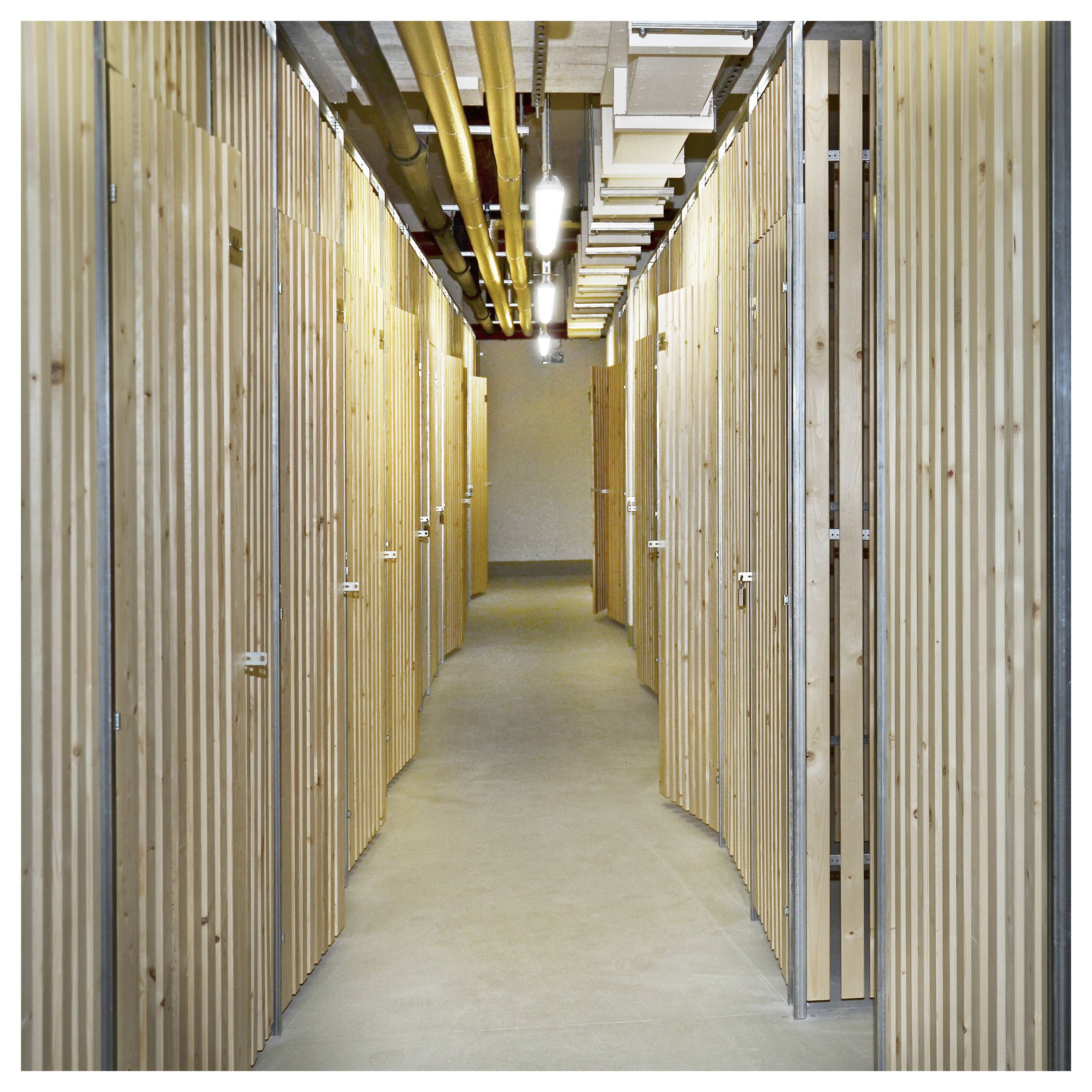 Ein Flur, gesäumt von Holzstauraum, Decke mit freiliegenden Rohren und Leuchten, der zu einer Endwand führt.