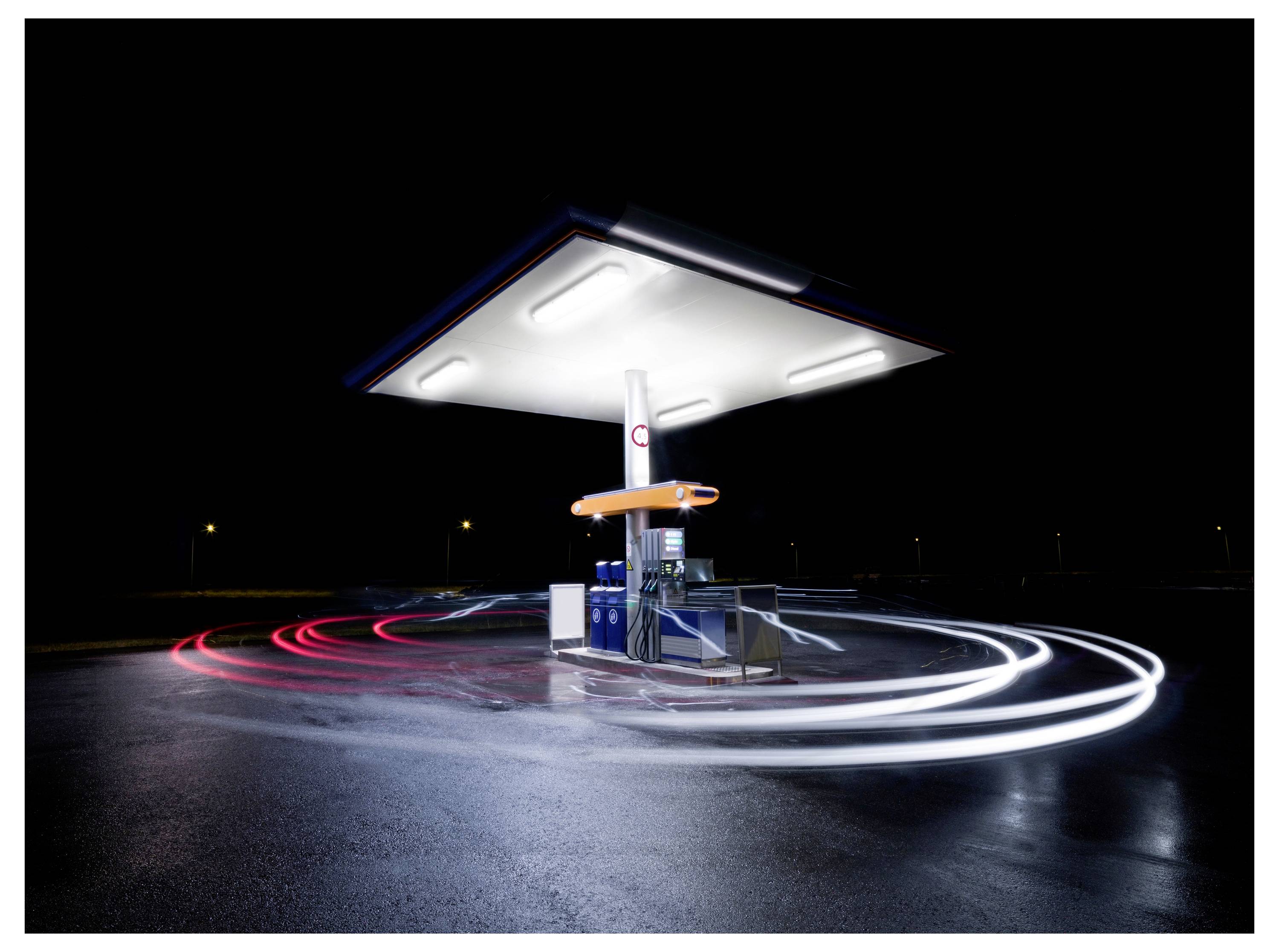 Eine hell erleuchtete Tankstelle bei Nacht, abgeschieden mit verschwommenen Lichtspuren um sie herum, die Bewegung und Aktivität andeuten.