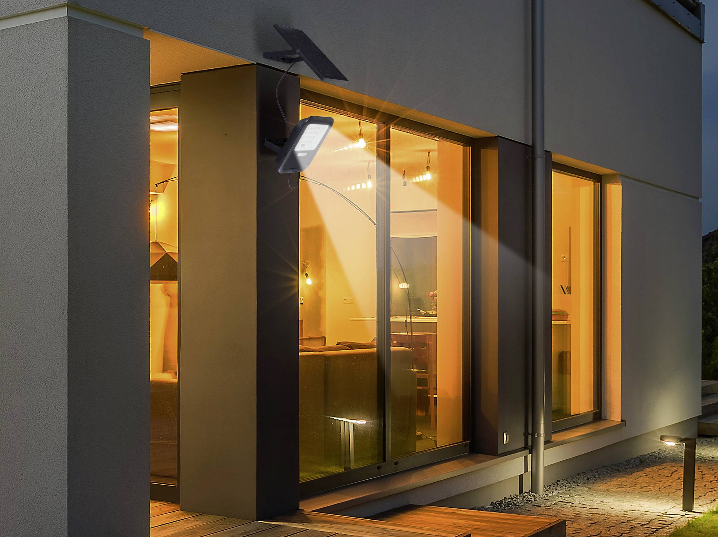 Solarlampe beleuchtet eine moderne Hausfassade bei Nacht. Große Fenster zeigen ein beleuchtetes Wohnzimmer.