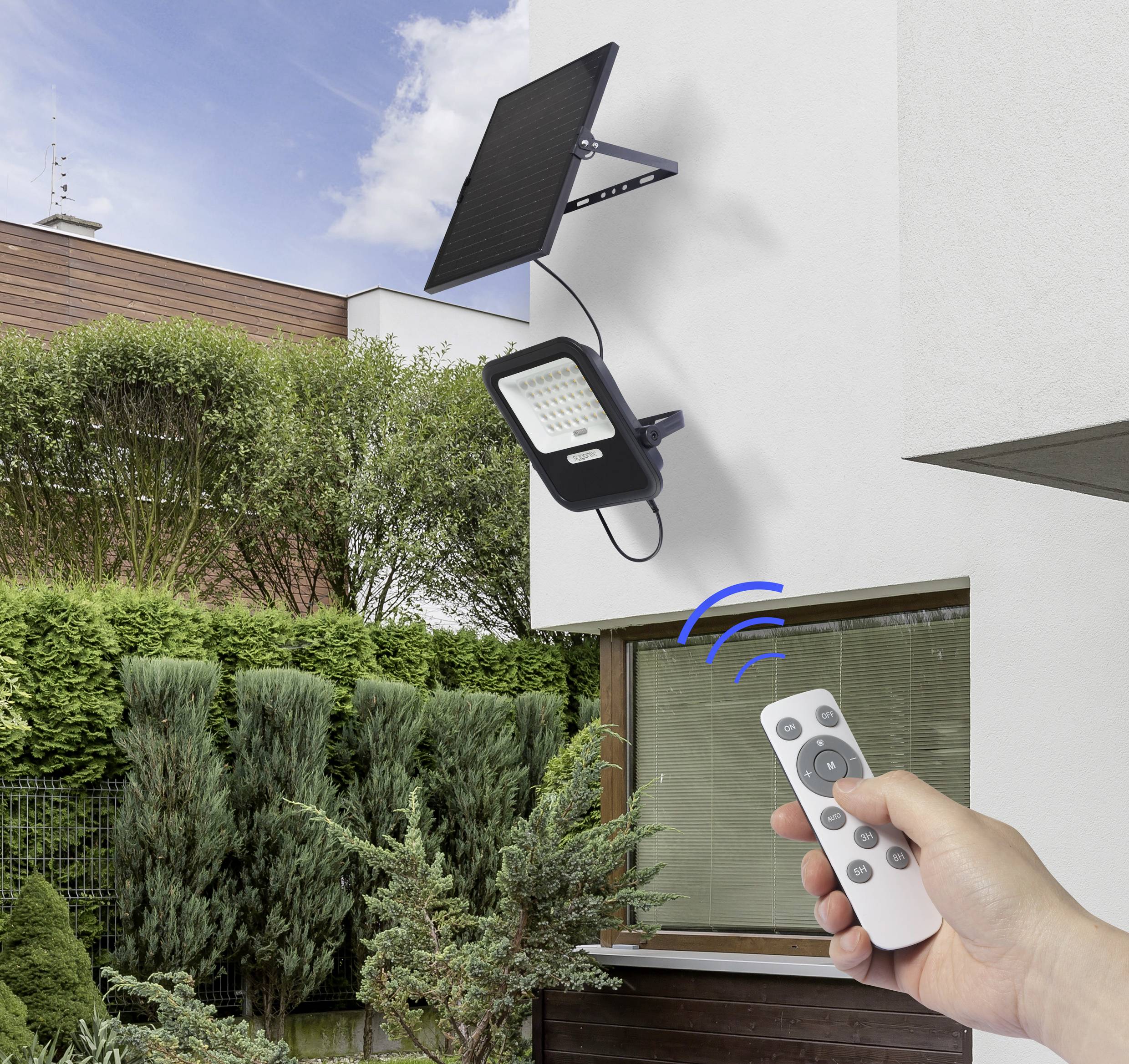 Eine Hand hält eine Fernbedienung, die eine solarbetriebene Außenleuchte an einem Haus aktiviert. Im Hintergrund Garten und blauer Himmel.