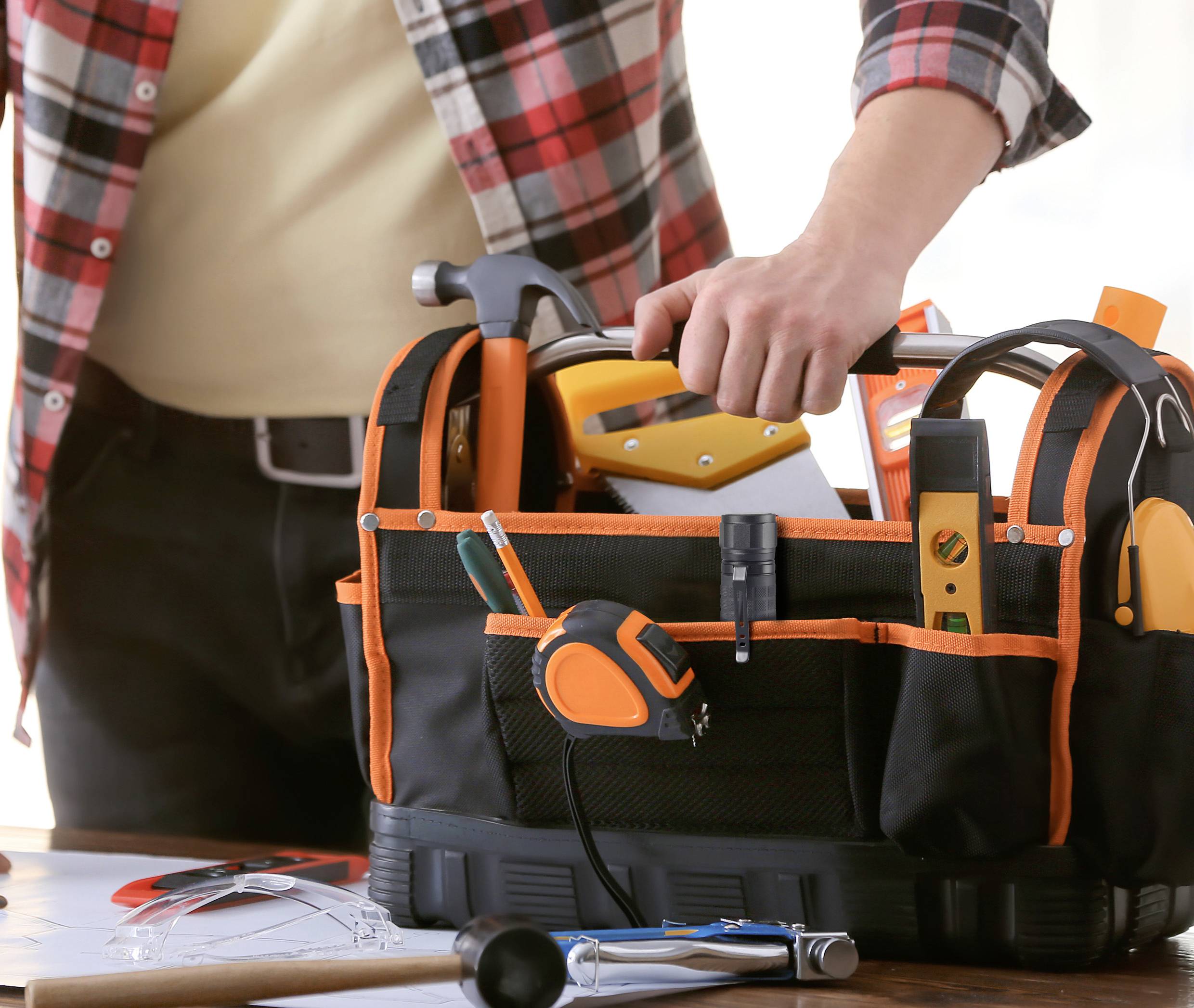 Person packt Werkzeuge in eine schwarze Werkzeugtasche mit orangefarbenen Details ein. Auf dem Tisch liegen Pläne und ein Hammer.