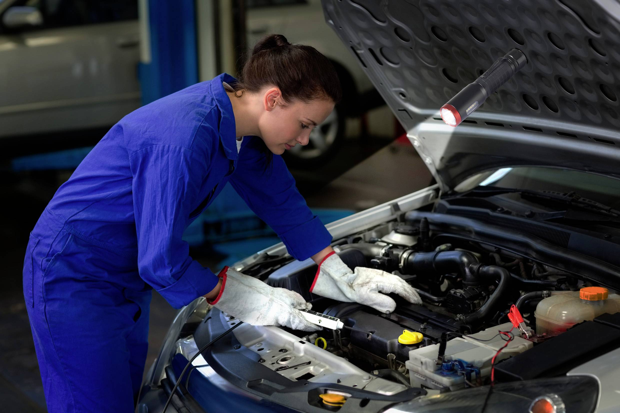 Person im blauen Overall untersucht den Motor eines Autos in einer Werkstatt.