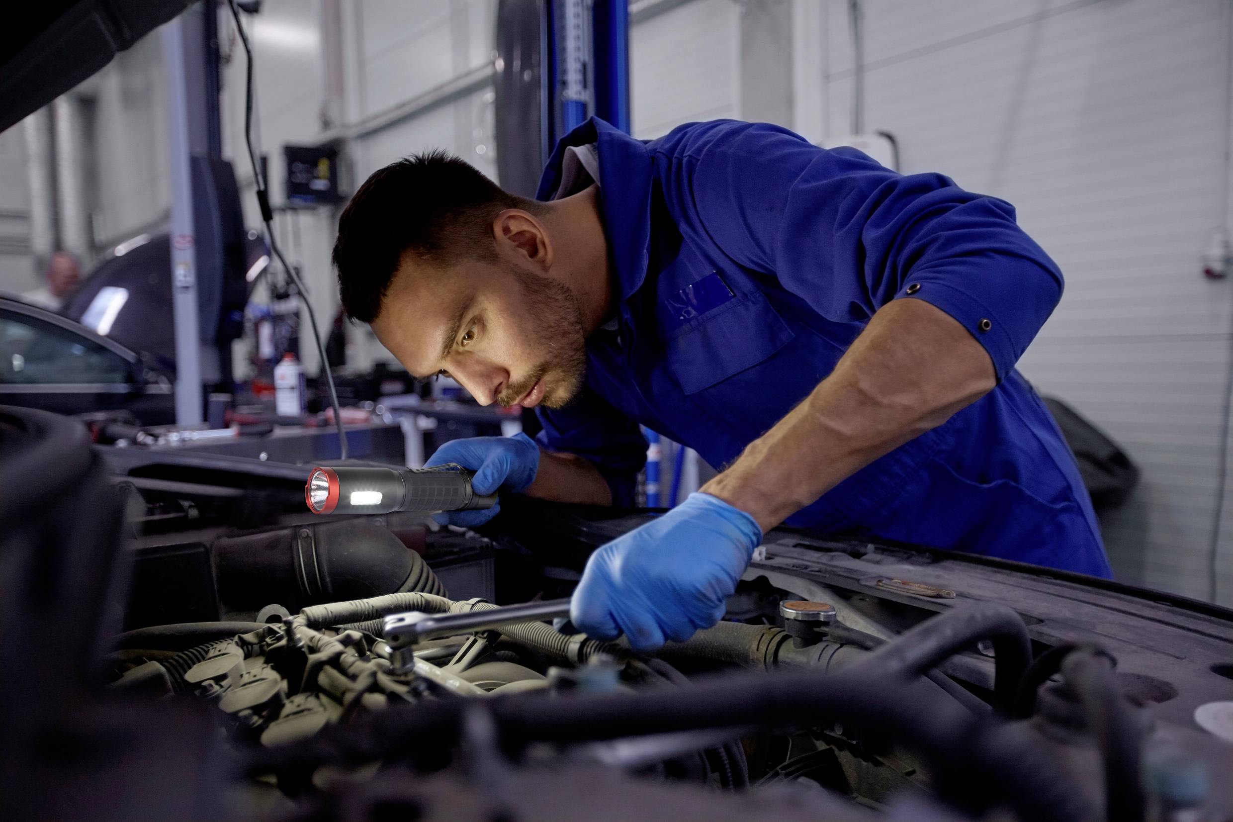 Ein Mechaniker in blauer Arbeitskleidung inspiziert mit einer Taschenlampe den Motor eines Autos in einer Werkstatt.