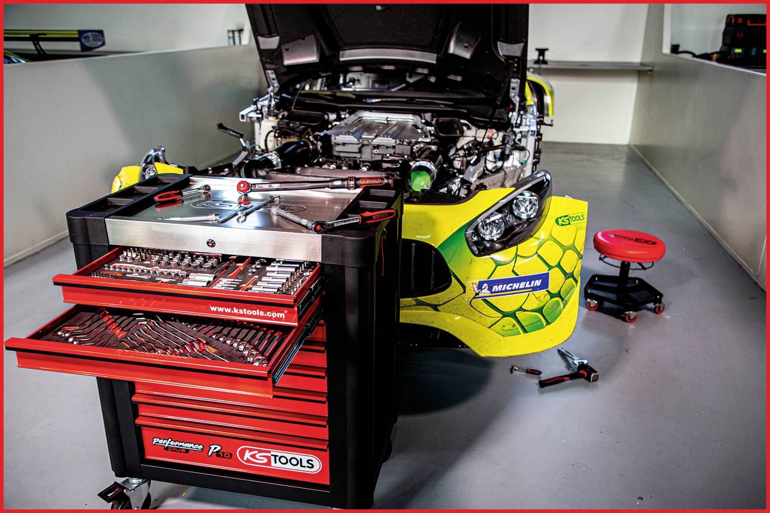 Renngarage mit offenem Sportwagen, Werkzeugwagen mit geöffneten Schubladen und verstellbarem Sitz. Im Hintergrund Werkstattumgebung.