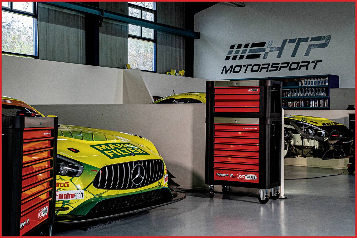 Ein grünes und gelbes Rennwagen in einer Werkstatt von 'HTP Motorsport' steht hinter roten Werkzeugschränken.