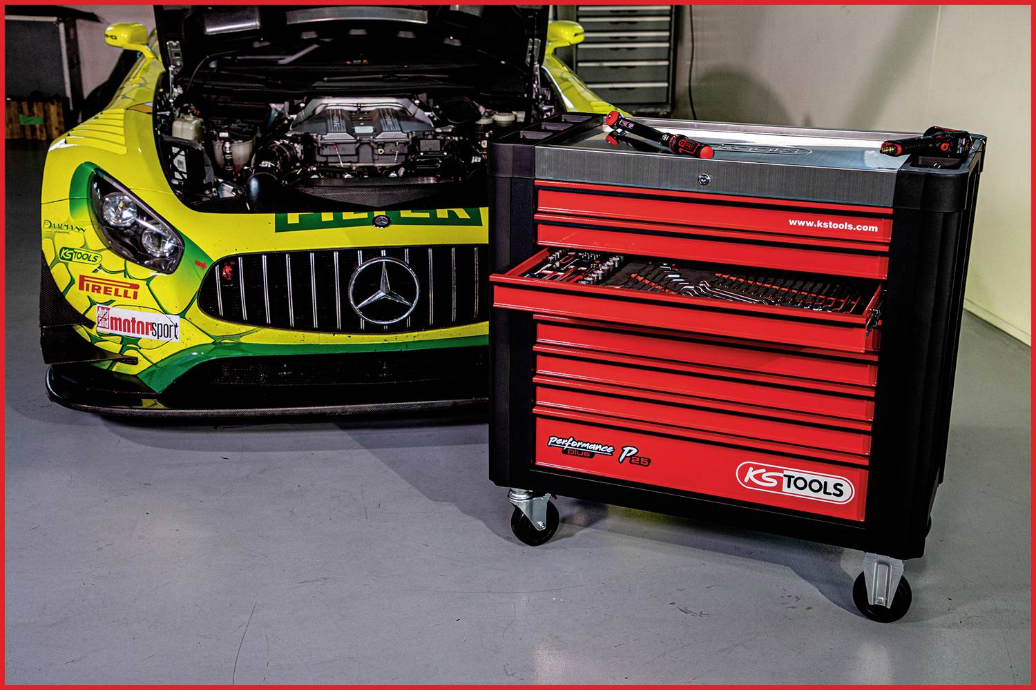 Ein Rennauto mit geöffnetem Motorraum und ein roter Werkzeugwagen voller Werkzeuge stehen in einer Werkstatt.