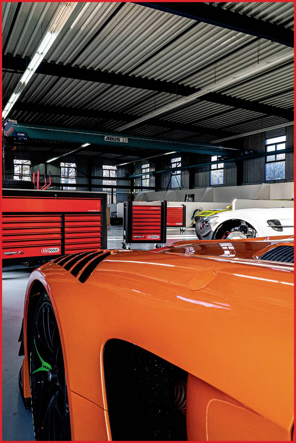 Ein orangefarbener Sportwagen steht in einer Werkstatt mit weiteren Fahrzeugen und Werkzeugschränken im Hintergrund.