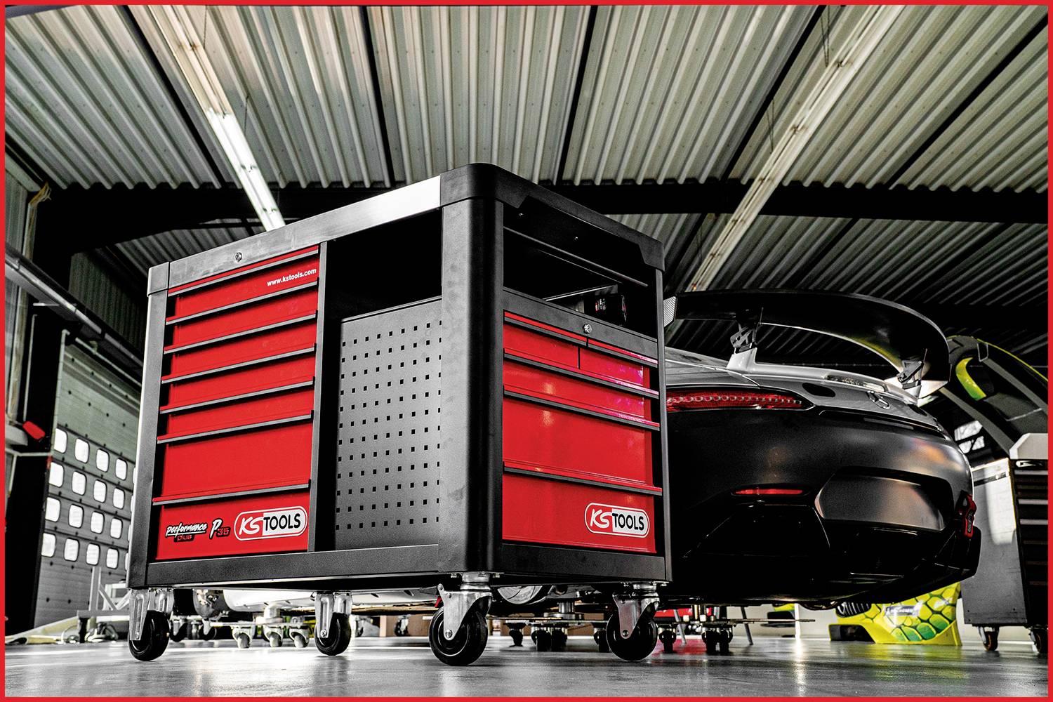 Werkstatt mit einem schwarzen Sportwagen, der in einer Garage geparkt ist. Im Vordergrund steht ein roter, rollbarer Werkzeugwagen.