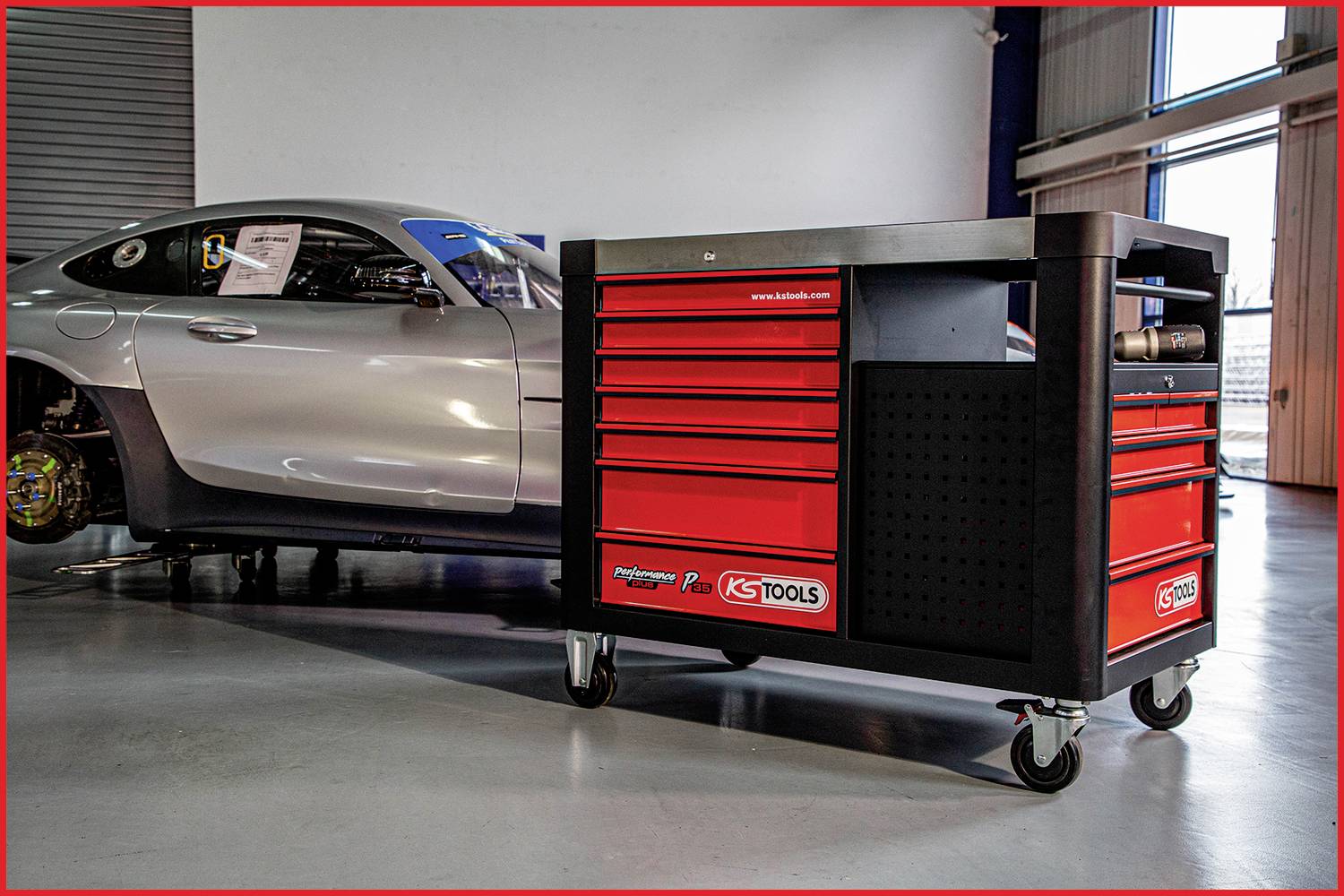 Ein silbernes Auto in einer Werkstatt, neben einem roten Werkzeugwagen auf Rollen, platziert auf einem grauen Boden.