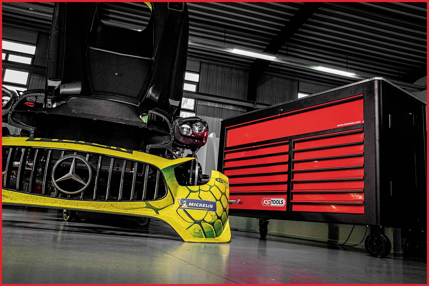 Ein gelber Rennwagenstoßfänger mit einem Schildkrötenmuster liegt neben einem roten Werkstattwagen mit Werkzeugschubladen in einer Garage.