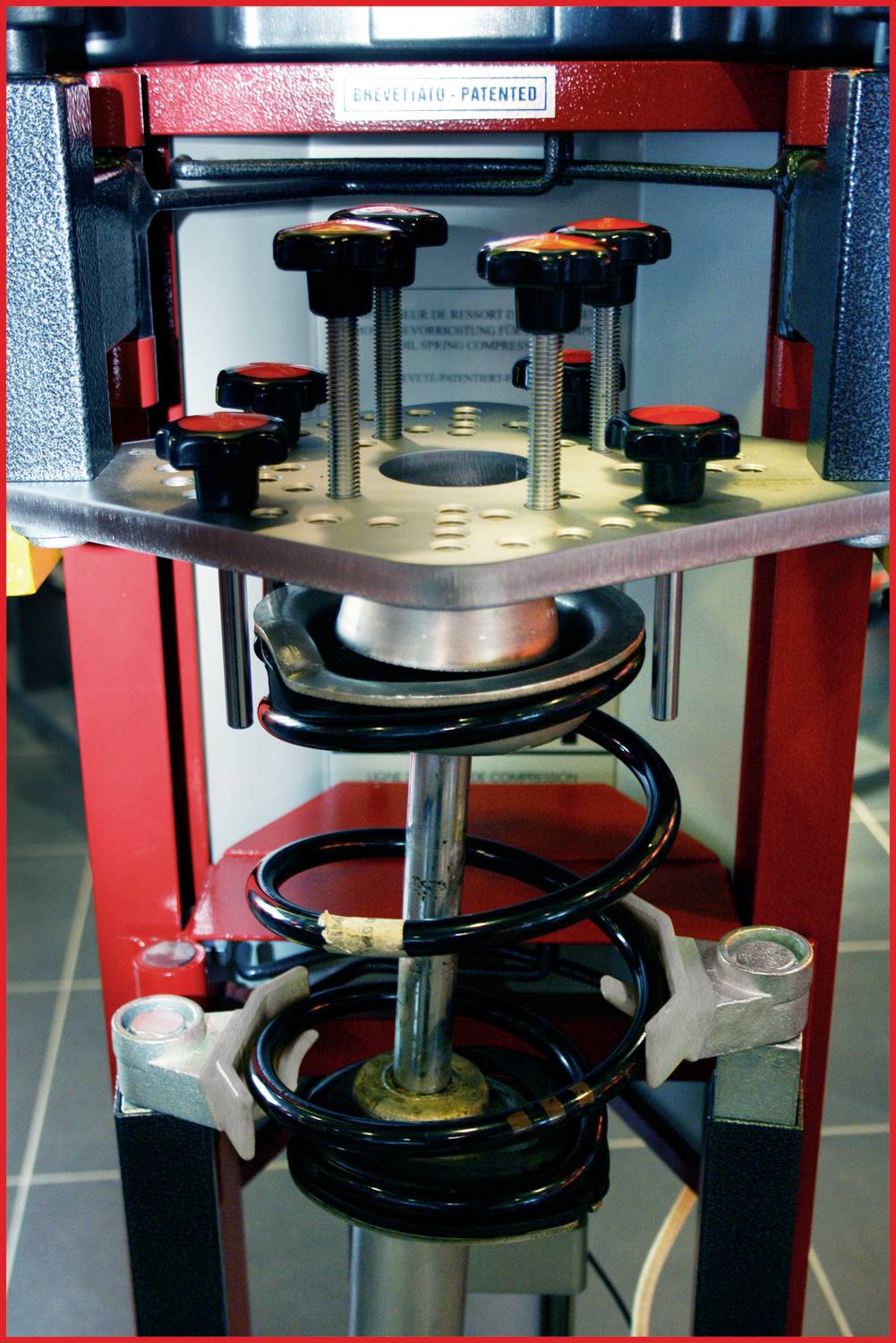 Eine rote Federpresse, die eine Metallfeder zusammendrückt. Die Schraubköpfe oben ermöglichen präzise Einstellungen.