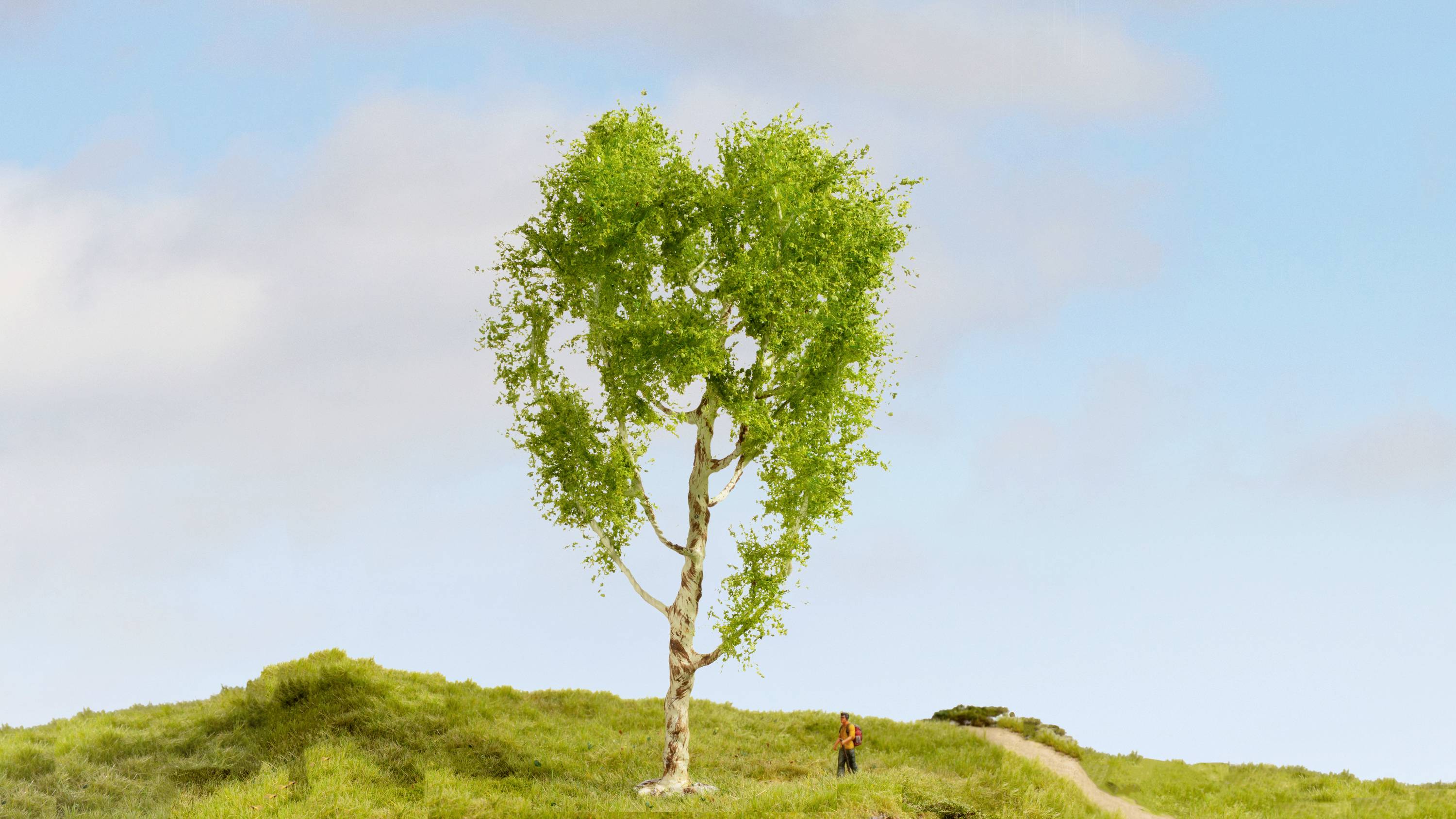 Ein Baum in Herzform steht allein auf einem grünen Hügel unter blauem Himmel. Eine kleine Person spaziert auf einem Pfad neben dem Baum.