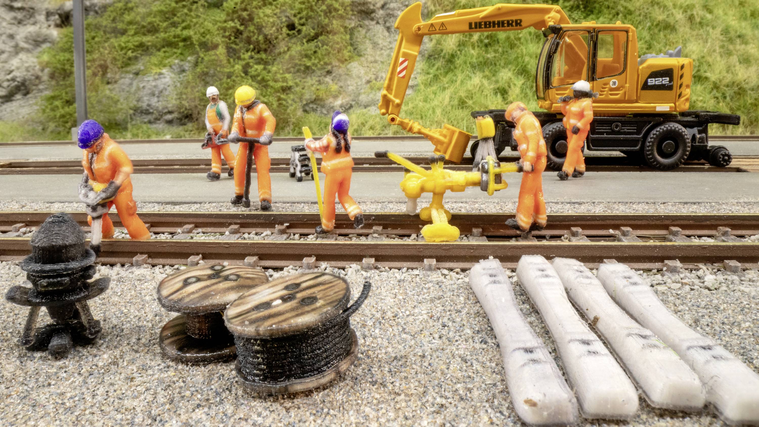 Arbeiter in orangefarbener Schutzkleidung reparieren Gleise. Im Hintergrund ein gelber Bagger. Im Vordergrund Kabelrollen und Schwellen.