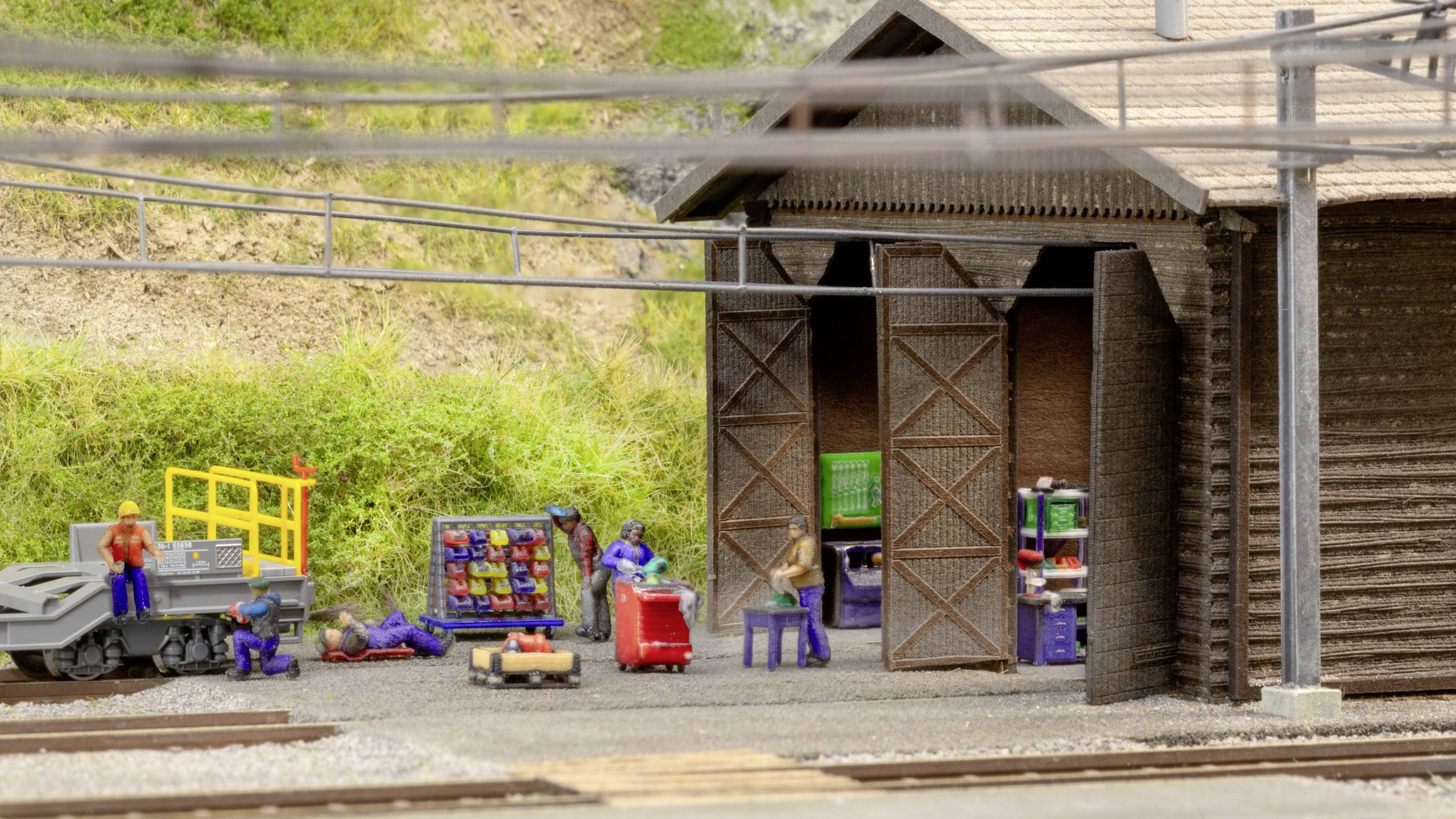 Ein Miniatur-Motorenwartungsbereich mit kleinen Figuren, die an einem Modellbahnwerkstattgebäude arbeiten. Kabel verlaufen darüber.