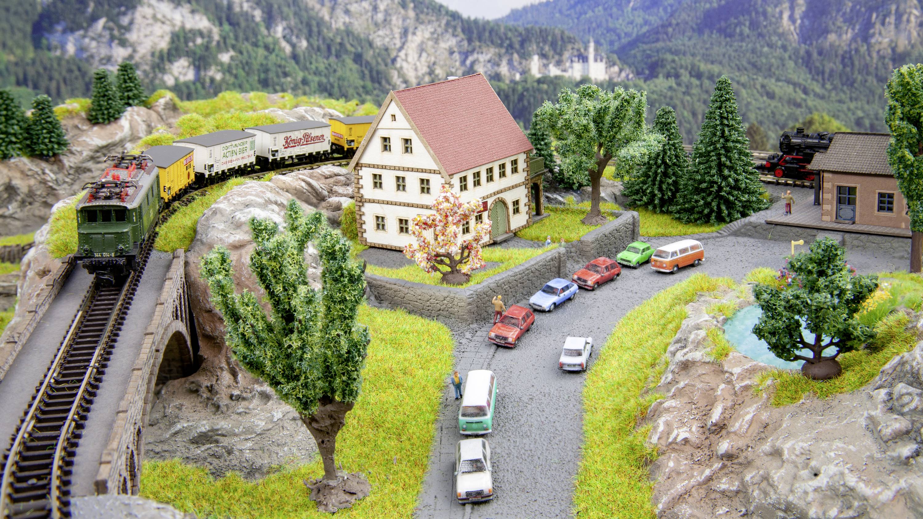 Eisenbahnmodelllandschaft mit Zug auf Brücke, mehreren Autos auf Straße und Gebäude umgeben von Bäumen, vor Bergkulisse.