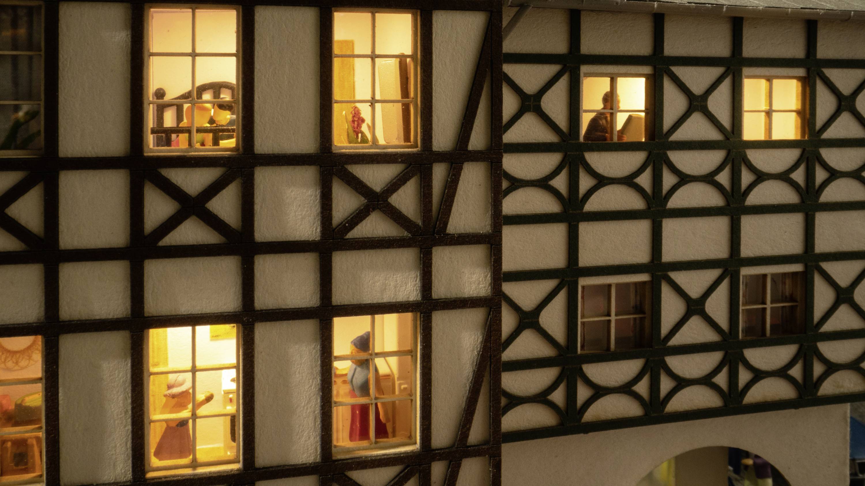 Fachwerkhäuser bei Nacht mit erleuchteten Fenstern, in denen Figuren zu sehen sind. Die Szene vermittelt ein ruhiges, warmes Ambiente.