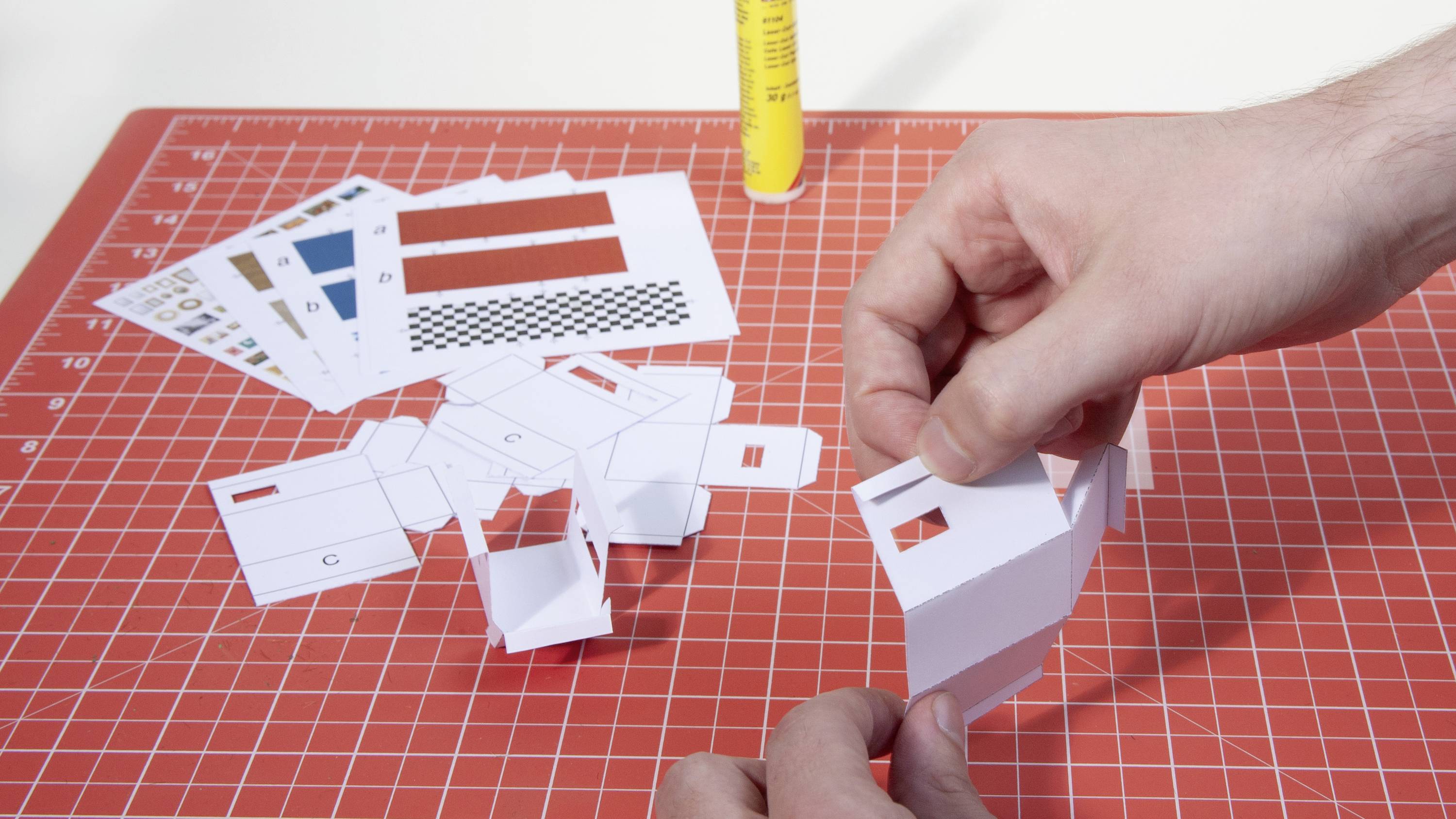 Eine Person faltet ein Modell aus Papier auf einer rot-weißen Schneidematte, umgeben von weiteren Papierstücken und Kleber.