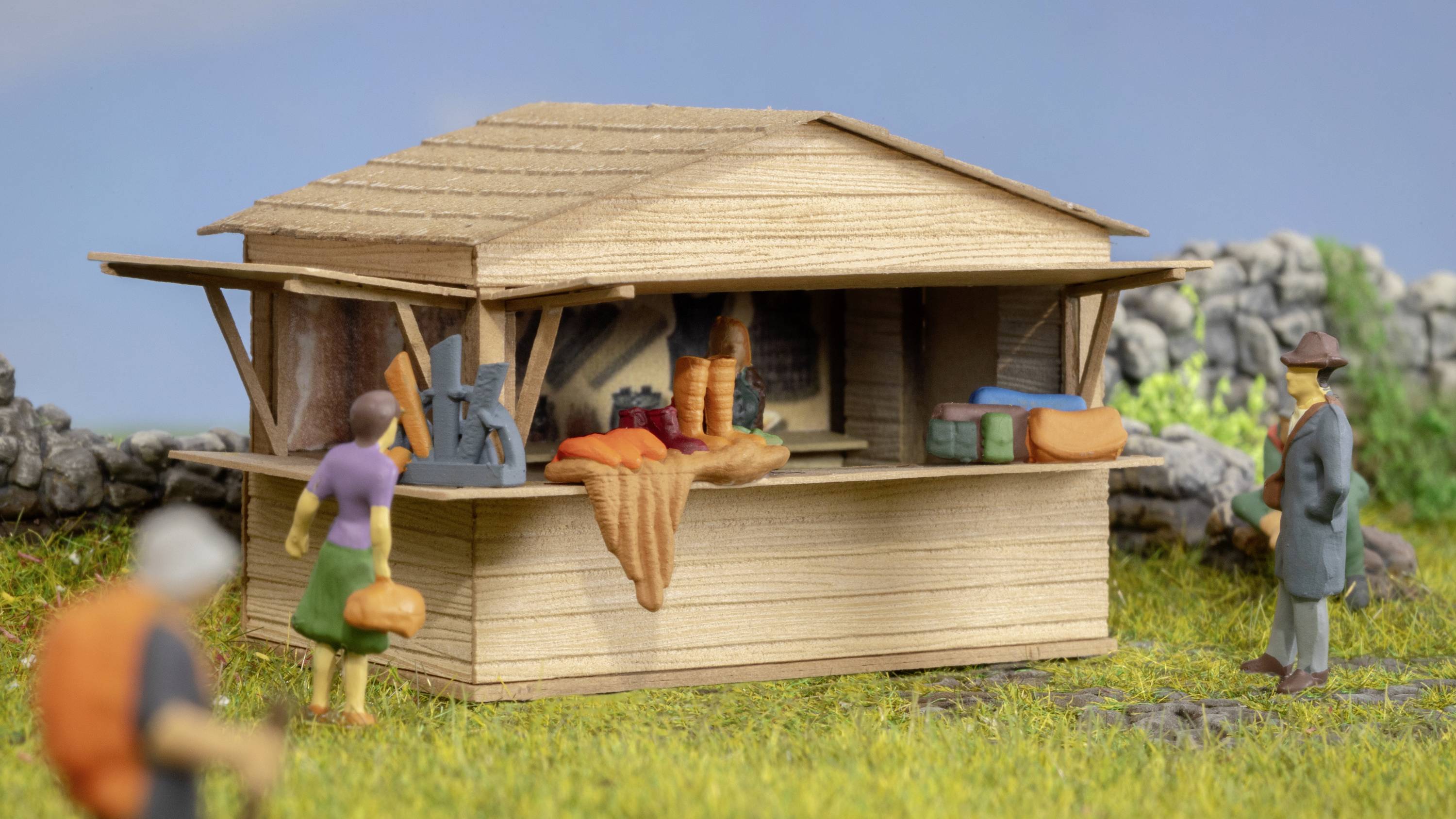 Ein kleiner Marktstand aus Holz mit diverser Ware wie Stoffe und Körbe. Zwei Figuren stehen davor, eine weitere Figur geht in der Nähe vorbei.