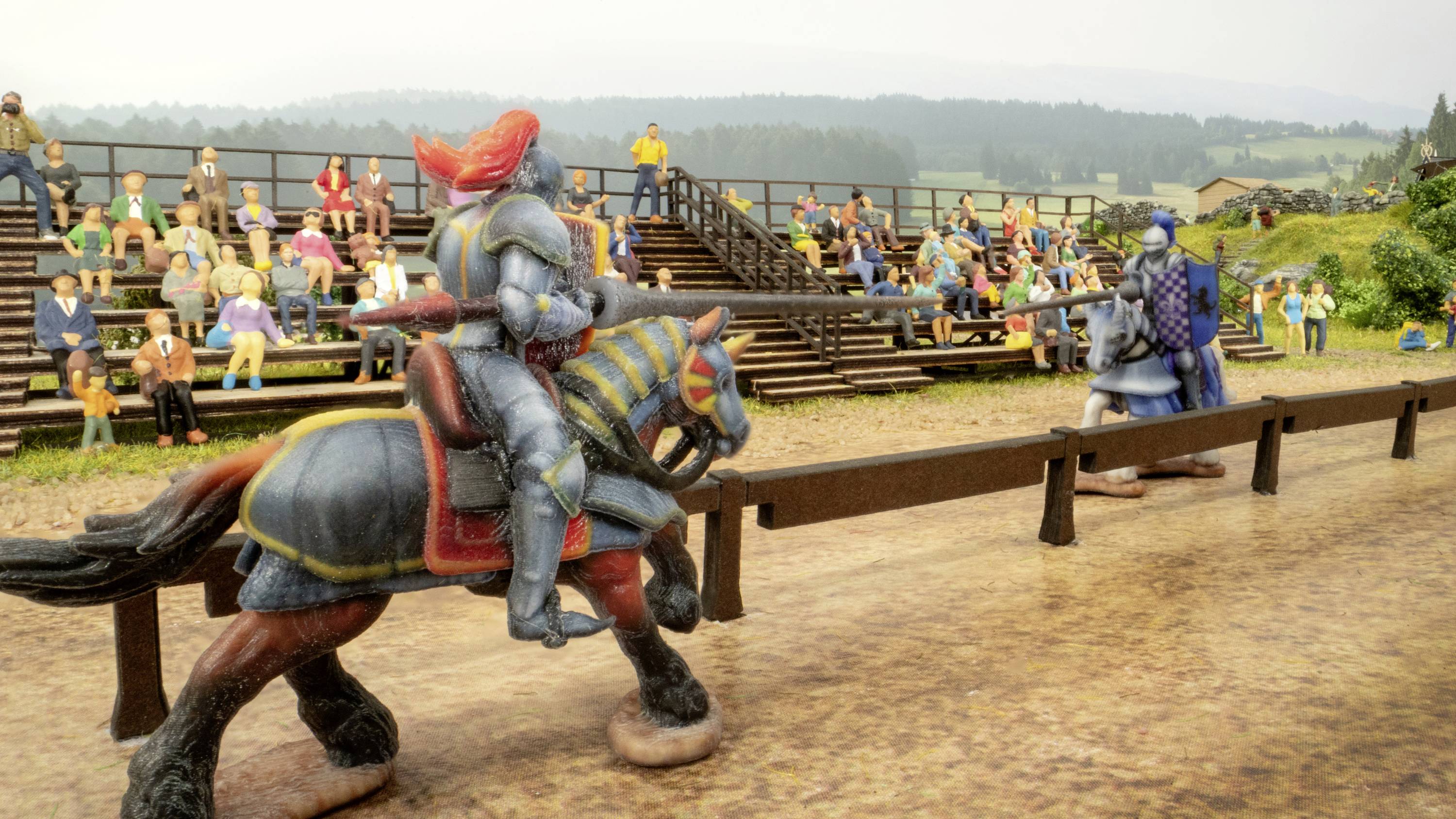 Zwei Ritter in Rüstungen zu Pferd bei einem Turnier. Zuschauer sitzen auf Tribünen im Hintergrund. Gebäude und Bäume in der Ferne.