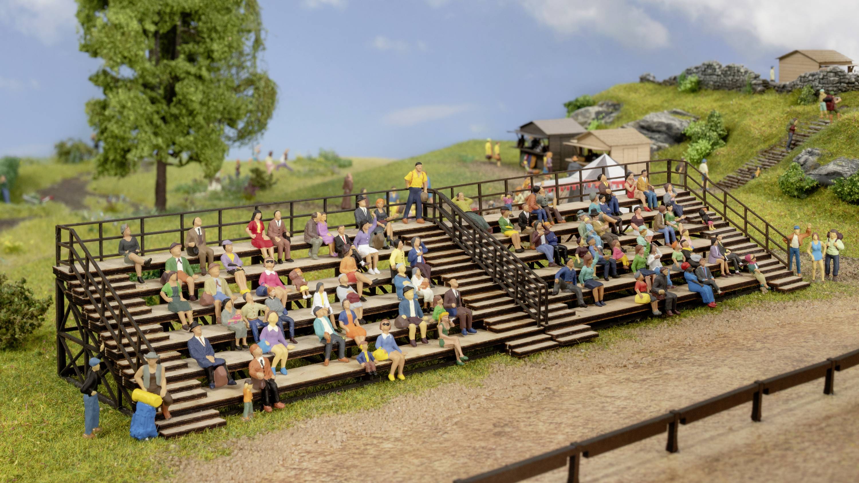 Miniaturmodell einer Tribüne im Freien mit zahlreichen sitzenden Zuschauern bei sonnigem Wetter, umgeben von Grünflächen und Bäumen.
