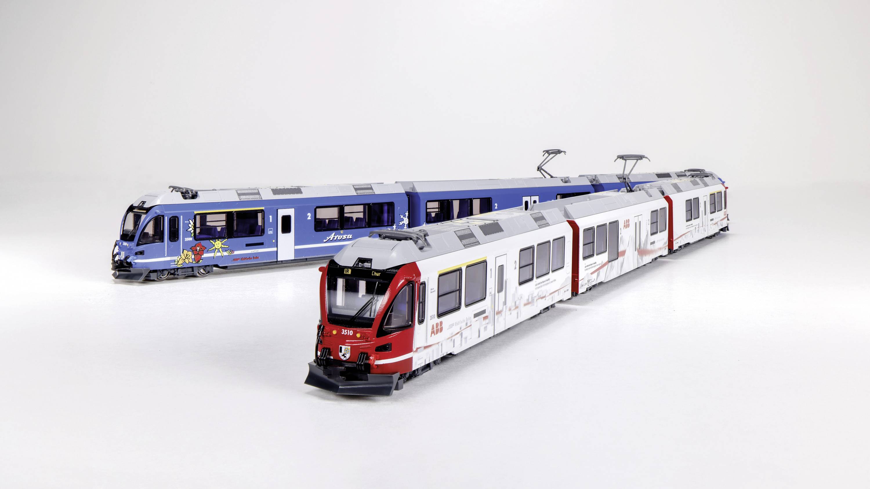 Zwei Modelleisenbahnen nebeneinander auf einer weißen Oberfläche: links ein blaues Modell mit buntem Logo, rechts ein weiß-rotes Modell.