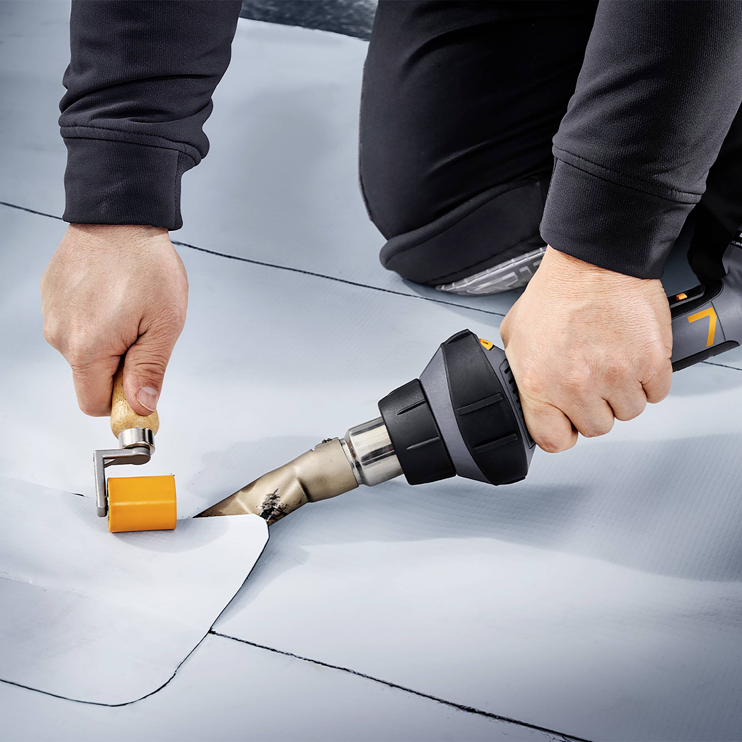 Zwei Hände arbeiten mit einem Handroller und einem Heißluftgebläse, um Schweißarbeiten an einer Folie durchzuführen.