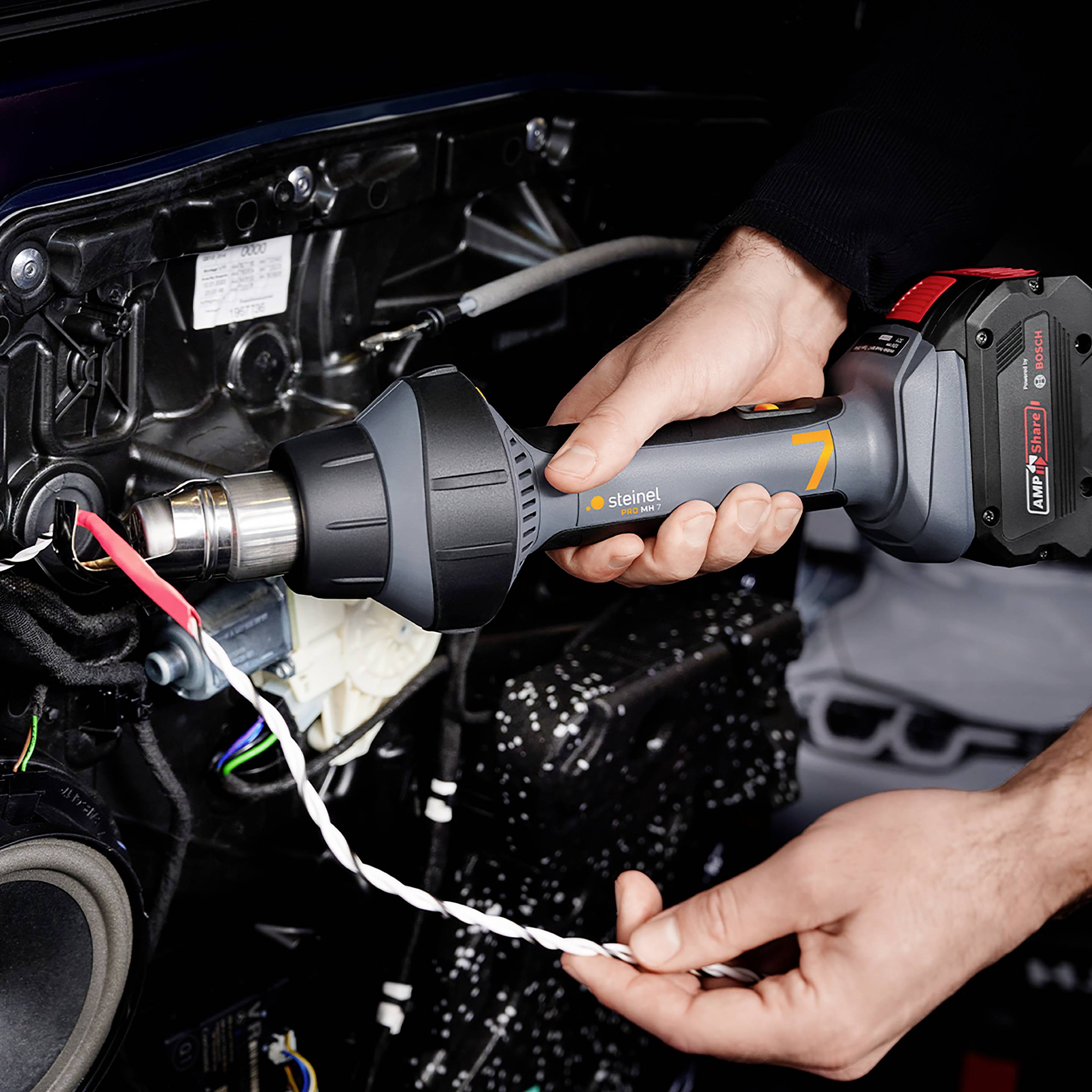 Eine Person bedient ein Heißluftgerät, das auf ein Kabel in einem Auto gerichtet ist. Hände halten das Gerät und das Kabel.