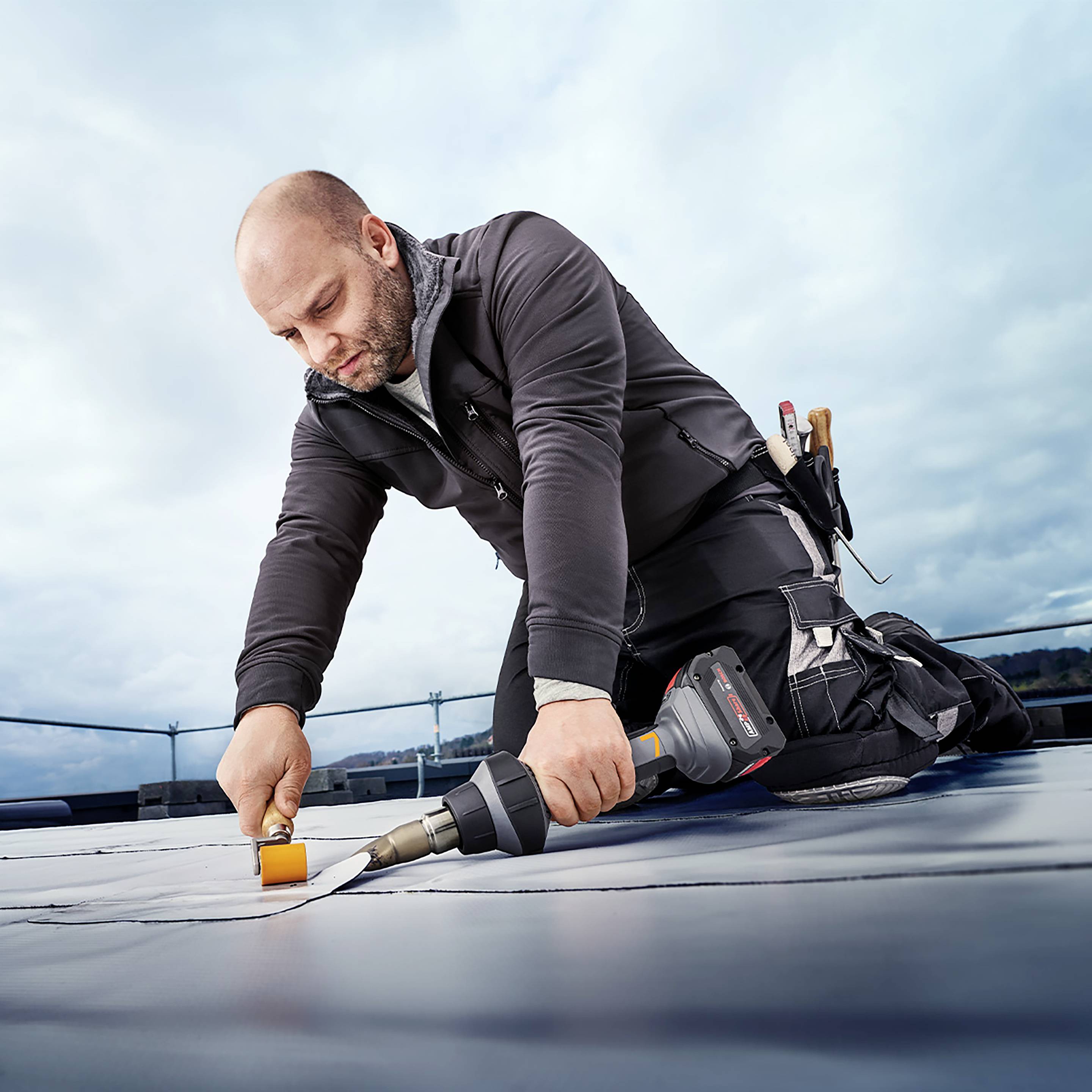 Ein Mann arbeitet auf einem Flachdach mit speziellem Werkzeug. Er trägt Arbeitskleidung und konzentriert sich auf die Aufgabe. Im Hintergrund ist der Himmel bewölkt.