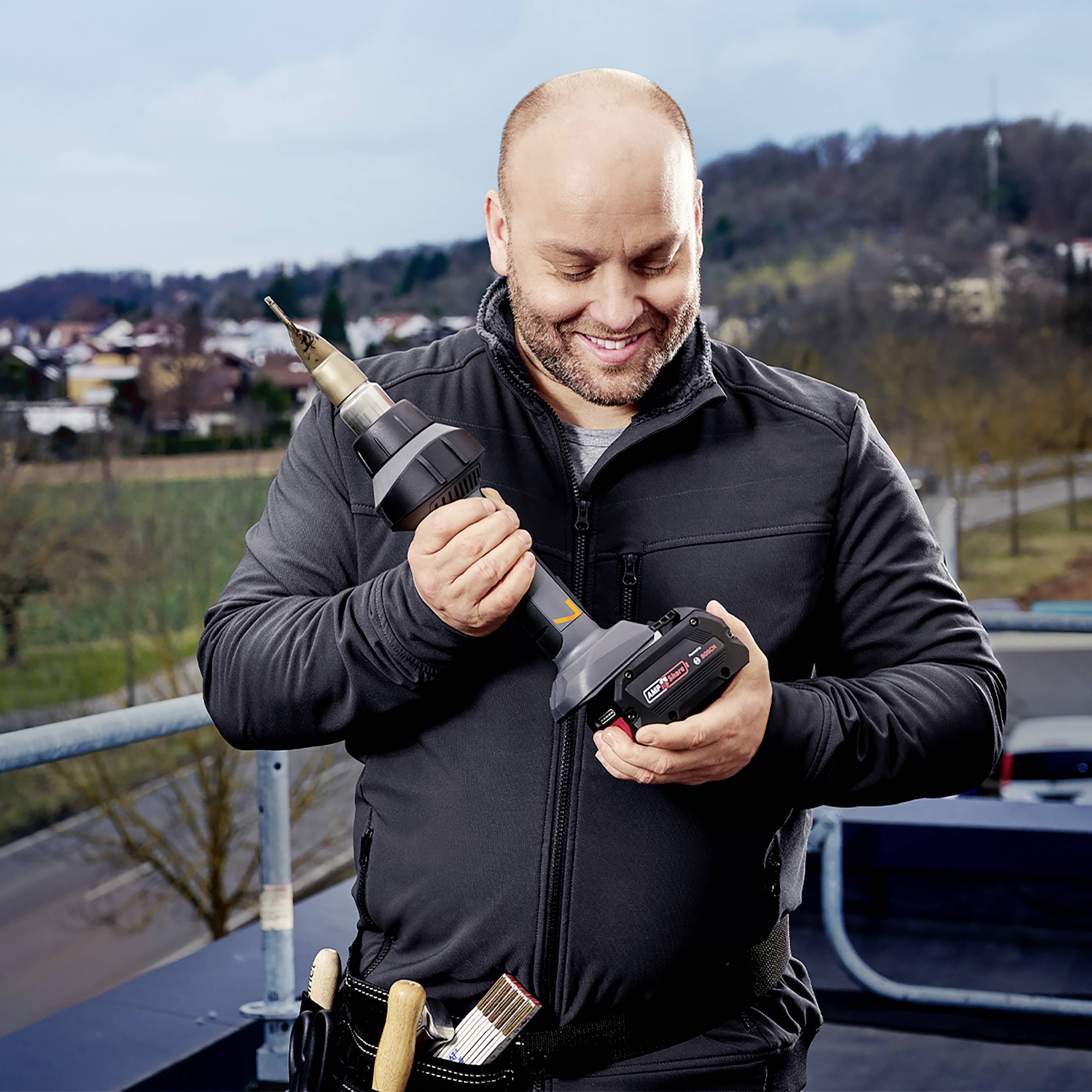 Eine Person auf einem Balkon hält eine Akkubohrmaschine und einen großen Bohrer, trägt Arbeitskleidung, lächelt und betrachtet das Werkzeug. Im Hintergrund eine Stadtlandschaft.