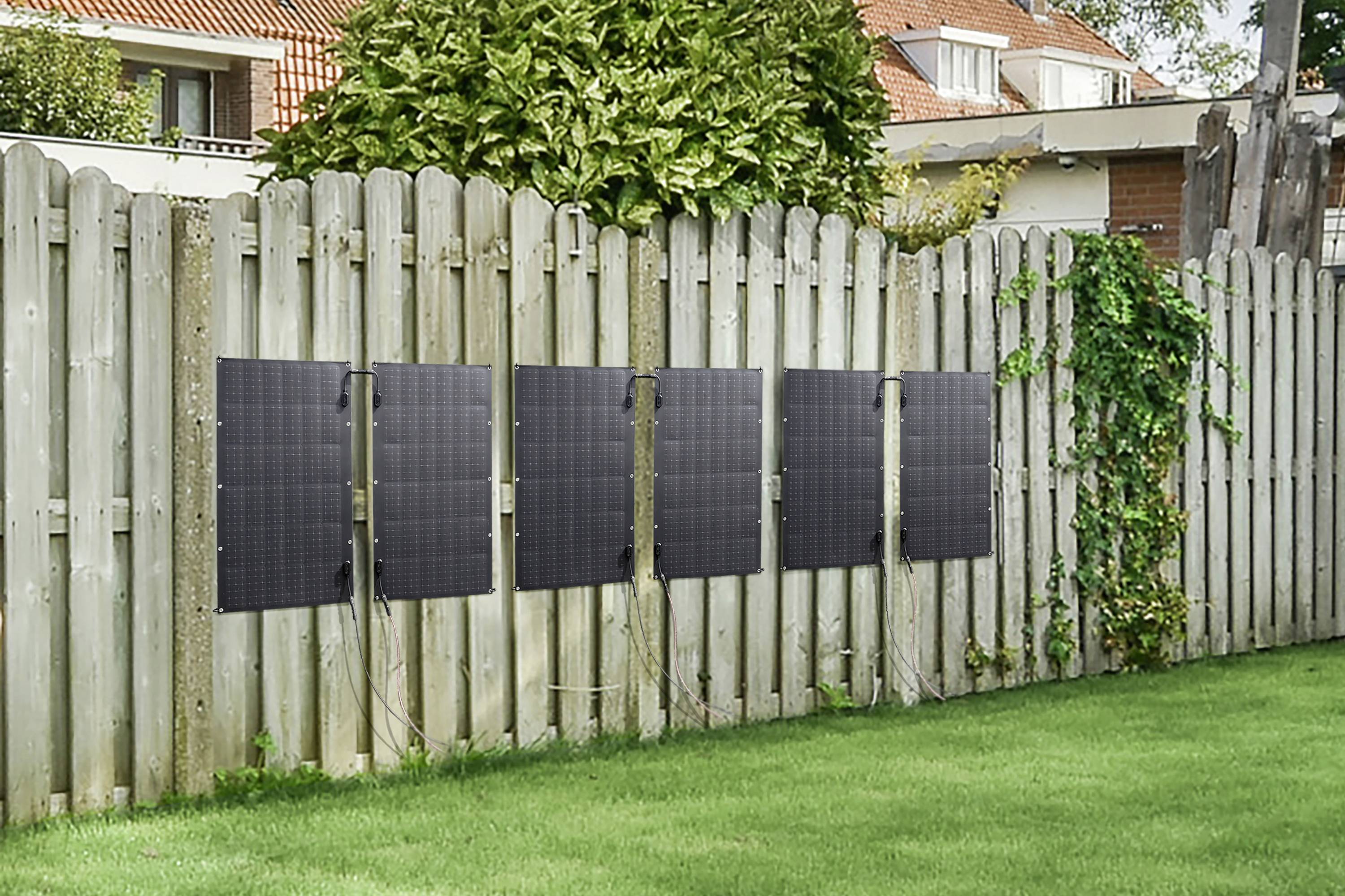 Fünf Solarmodule sind an einem Holzzaun im Garten montiert. Im Hintergrund sind Häuser und Bäume zu sehen.