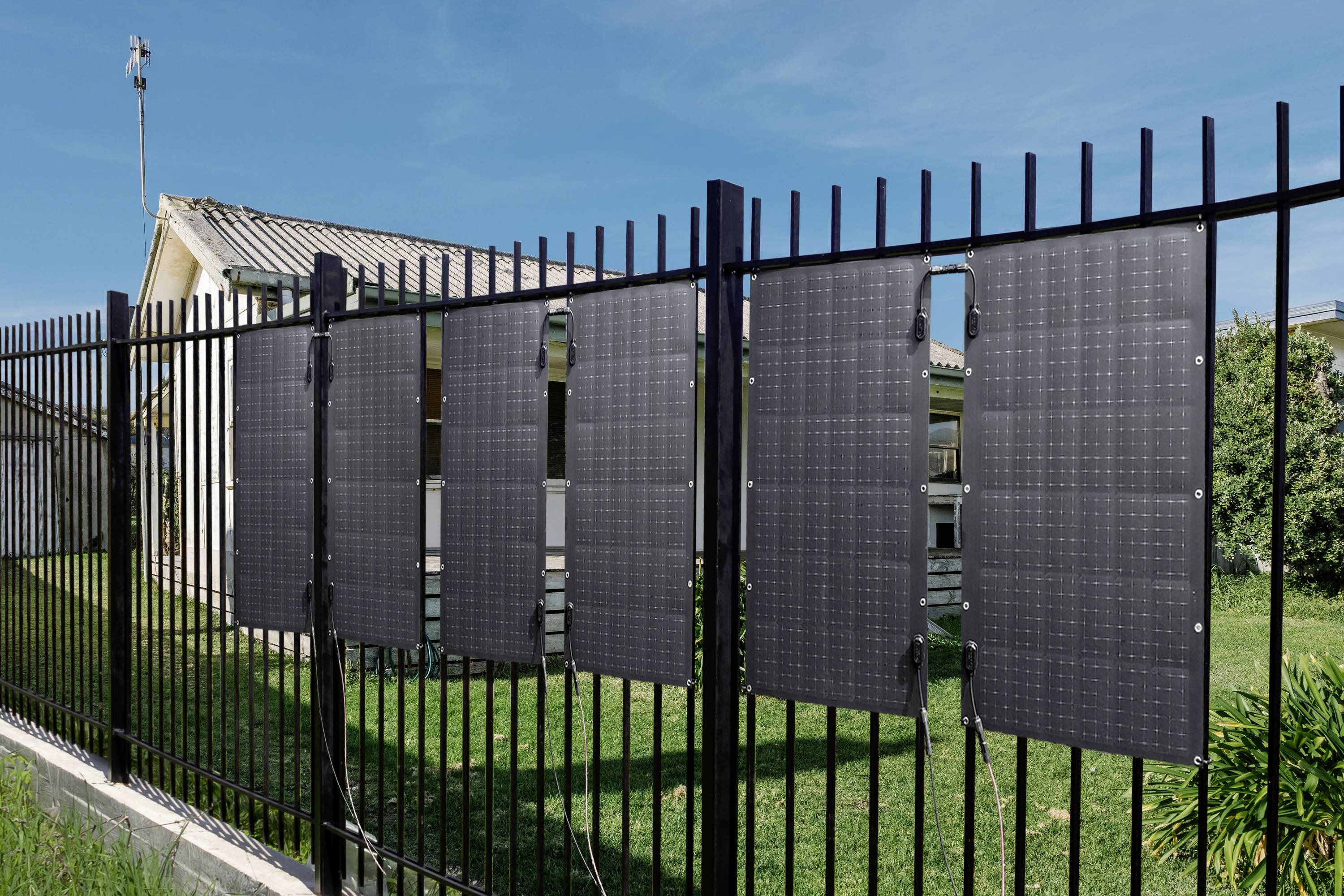 Solarmodule sind an einem Metallzaun befestigt, mit einem Haus im Hintergrund. Die Szene zeigt nachhaltige Energienutzung in einem Wohngebiet.