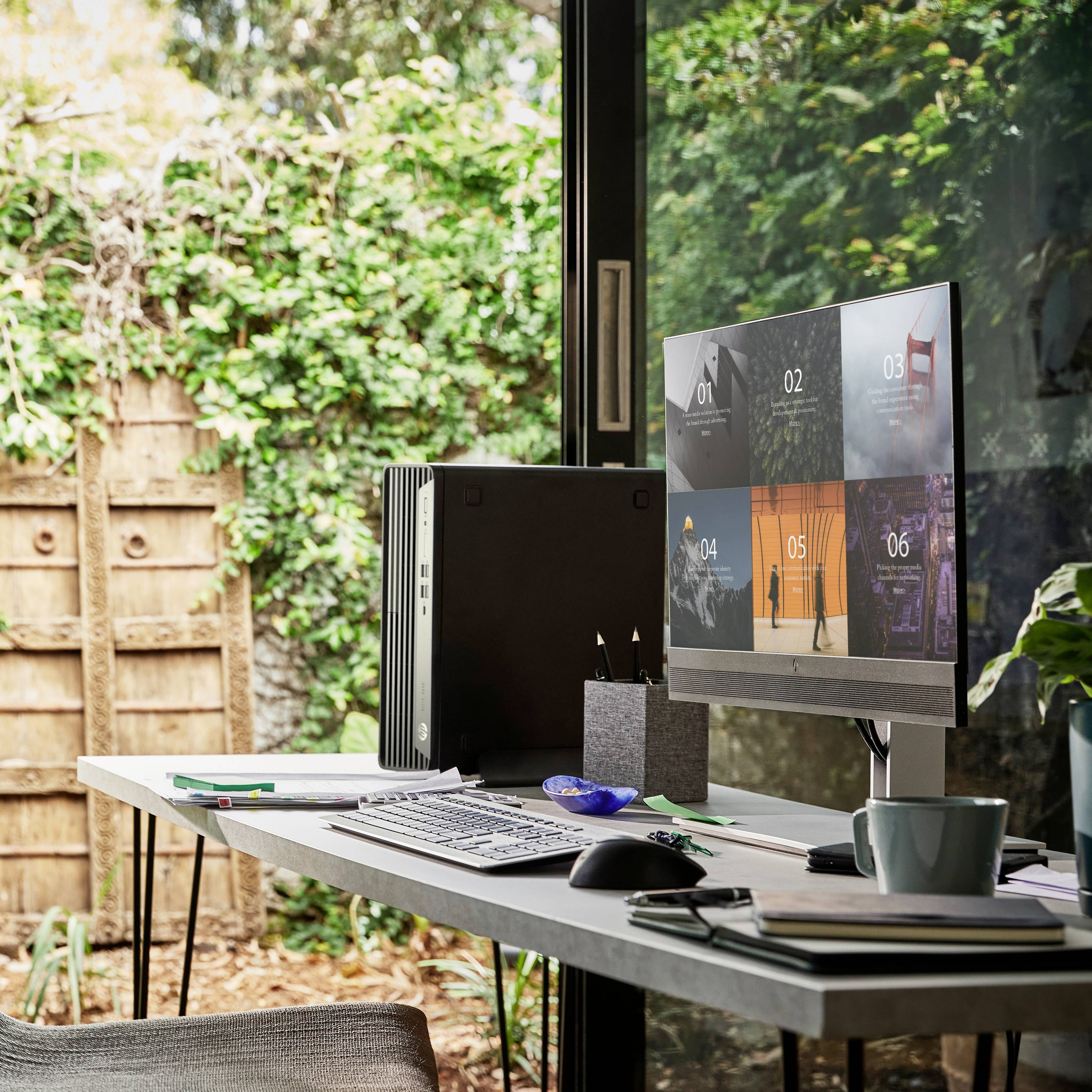 Schreibtisch am Fenster mit Computer, Tastatur, und Notizbuch, Blick nach draußen auf Garten mit Holzzaun und Pflanzen.